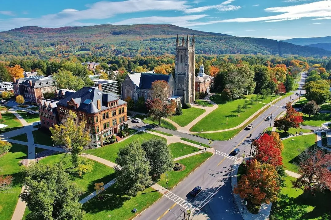 An aerial view of a college campus surrounded by colorful trees in autumn. The scene includes a church steeple and a mix of historical and modern buildings.