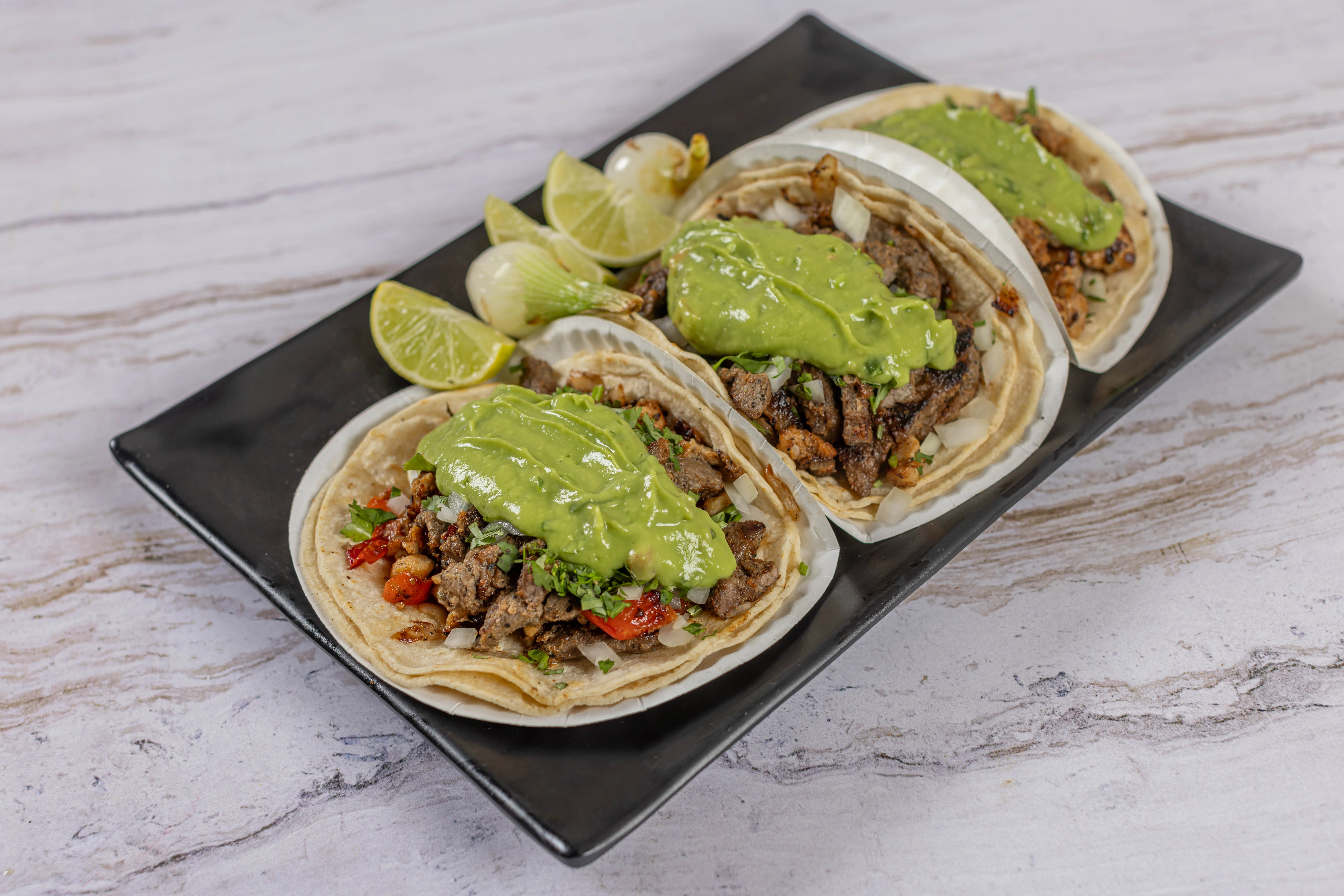 A plate of three authentic steak tacos served on corn tortillas with creamy avocado salsa, grilled onions, and lime.