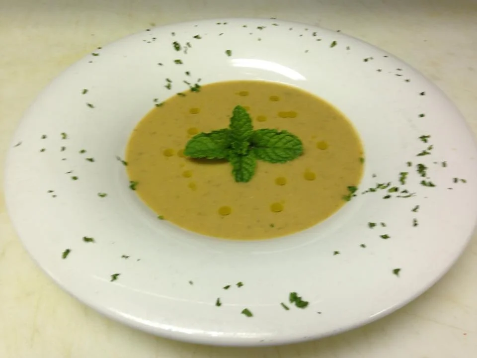 A bowl of creamy soup garnished with fresh mint leaves and dotted with olive oil, served on a white plate.