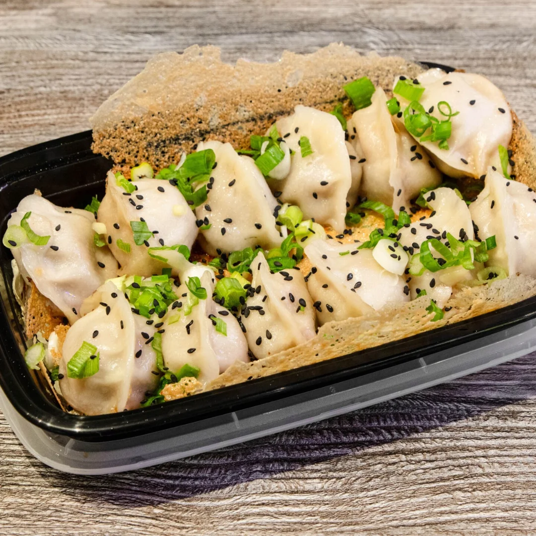 Steamed dumplings topped with green onions and sesame seeds in a black container.