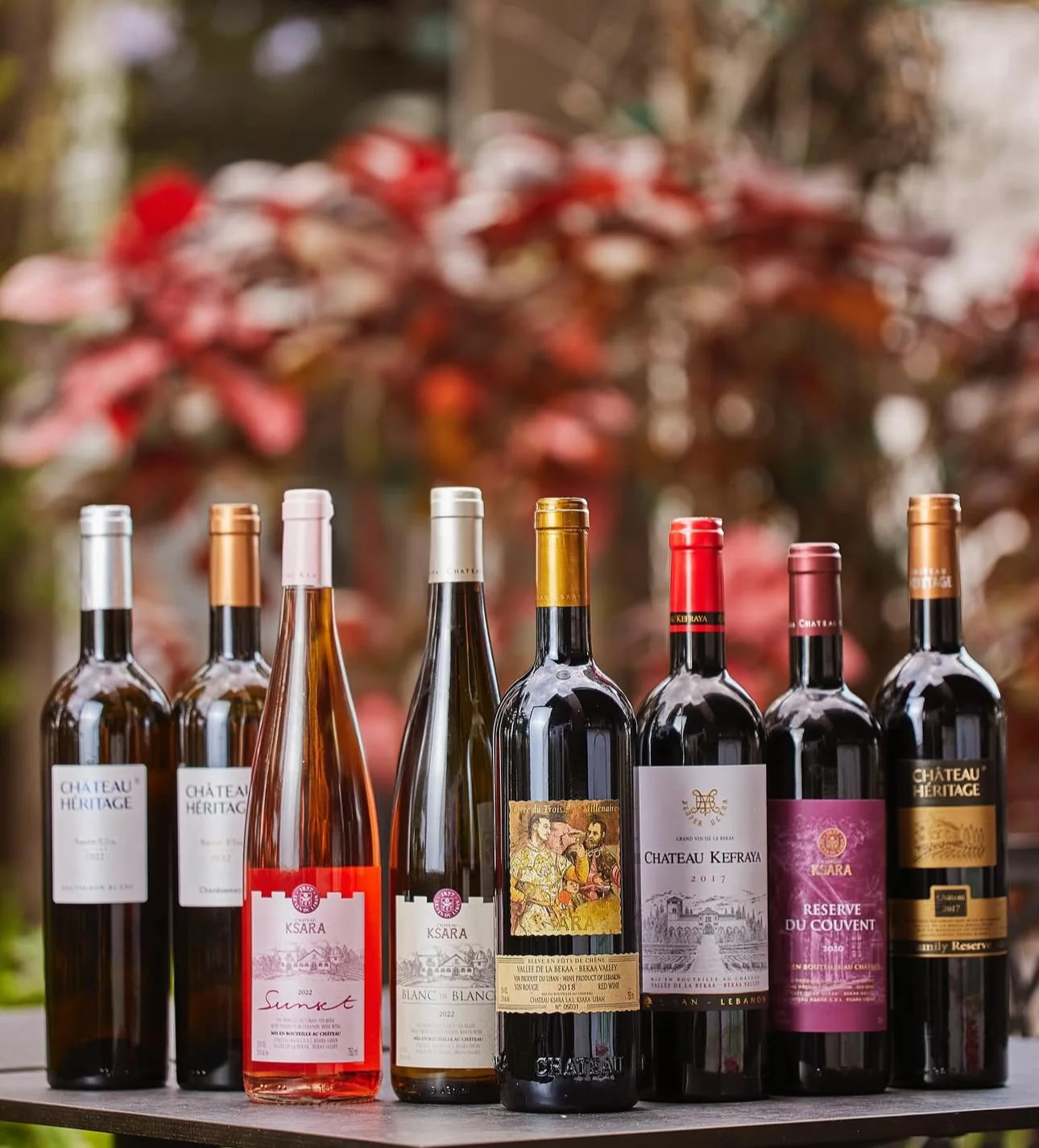 A row of eight assorted wine bottles, with colorful labels, stands against a blurred backdrop of vibrant autumn leaves. The scene conveys a warm, inviting atmosphere.