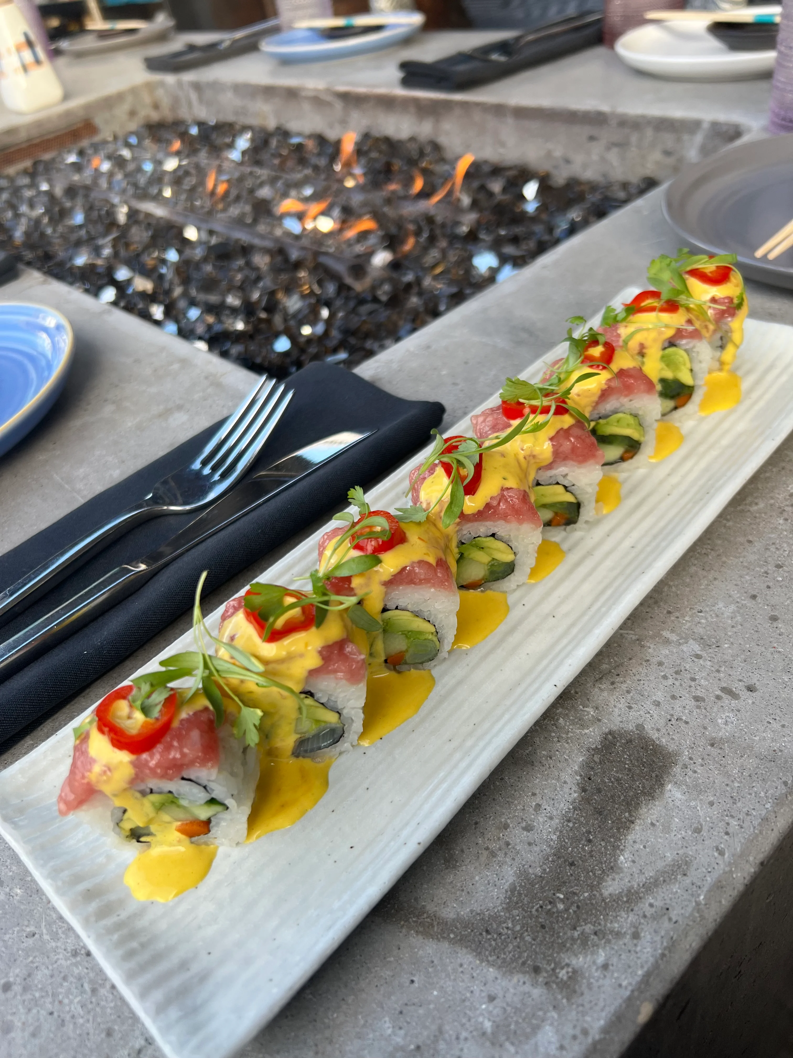 A beautifully presented plate of sushi with avocado, tuna, and a yellow sauce, garnished with red chili and herbs, placed on a granite tabletop with a fire feature in the background.