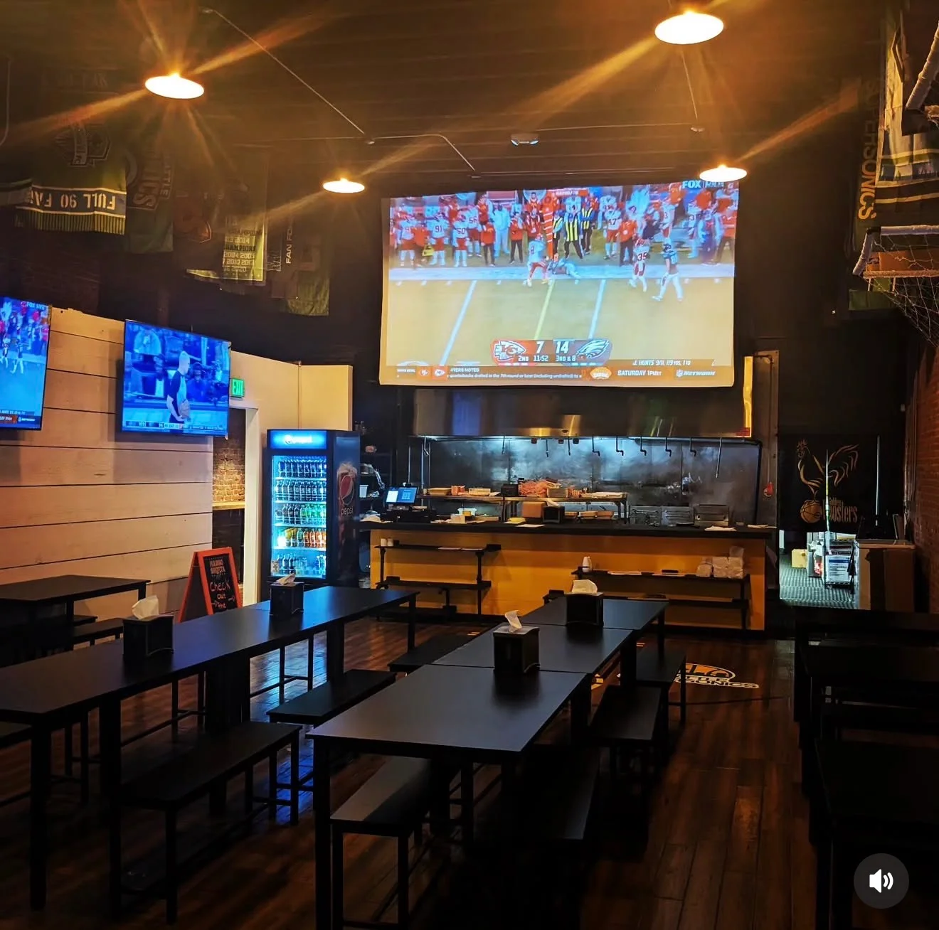 An indoor sports bar setting with multiple tables and a large screen displaying a sports game.