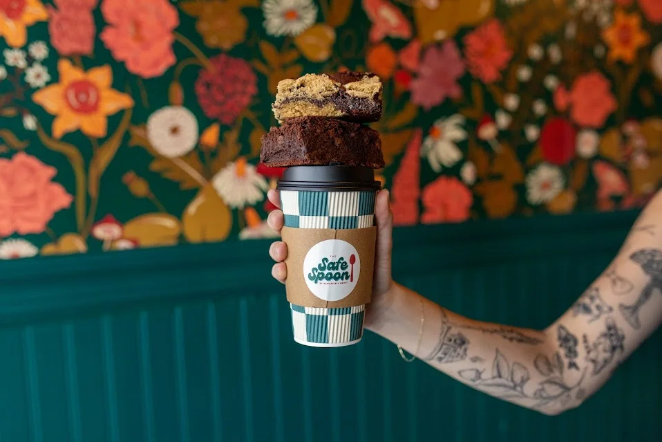 A hand holds a coffee cup topped with two pieces of dessert, set against a vibrant floral backdrop.