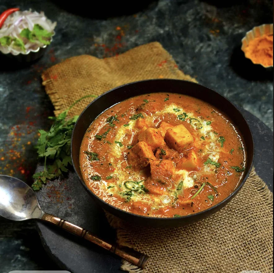 A spicy paneer curry garnished with cream and herbs, served in a black bowl on a textured background.