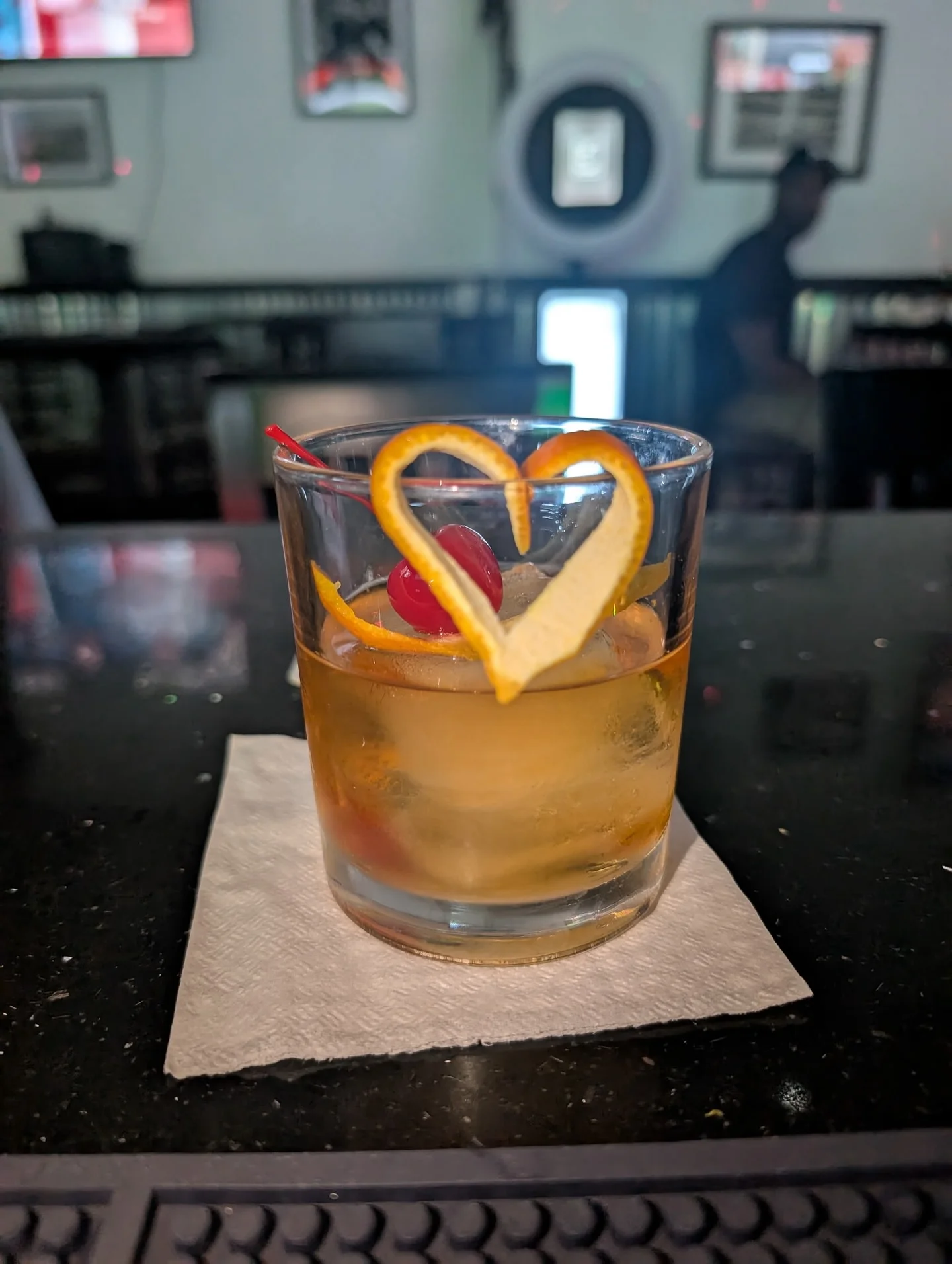 A cocktail in a glass garnished with a heart-shaped orange peel and a cherry, placed on a napkin.