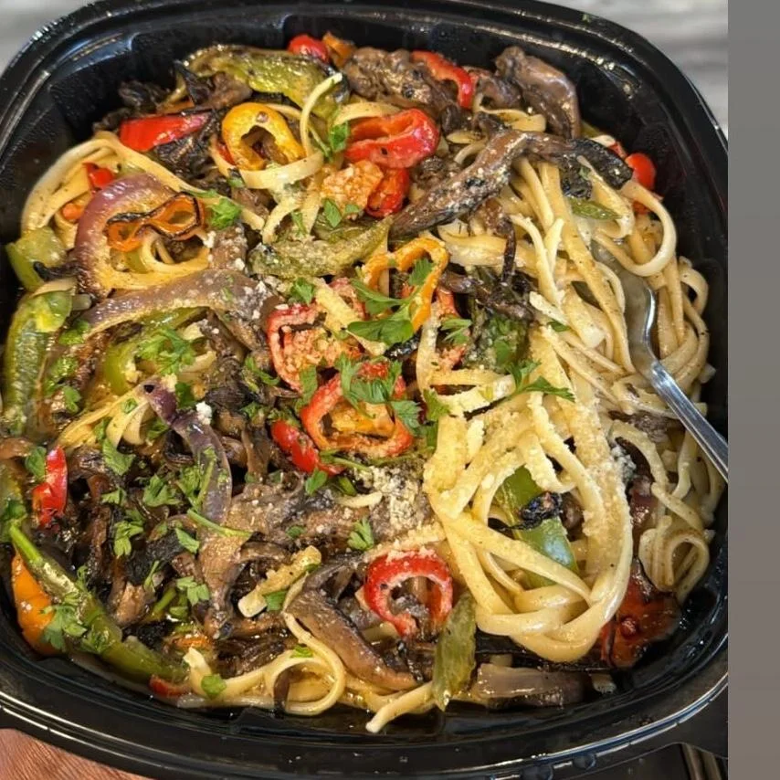 A bowl of pasta with mixed vegetables, garnished with herbs and cheese, presented in a black container.