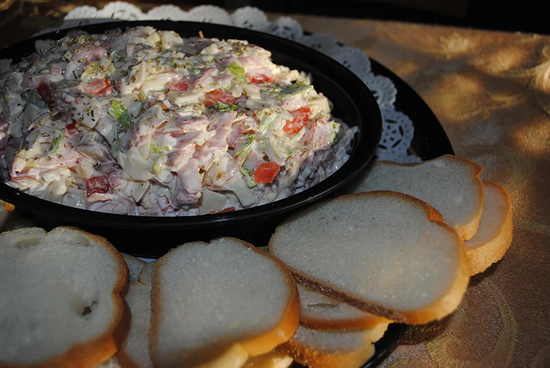 A black dish filled with a creamy salad mixture containing diced ham, diced salami, vegetables, and garnished with seasoning, and surrounded by slices of white bread on a black platter with a decorative lace doily. The platter is placed on a table with a golden tablecloth.