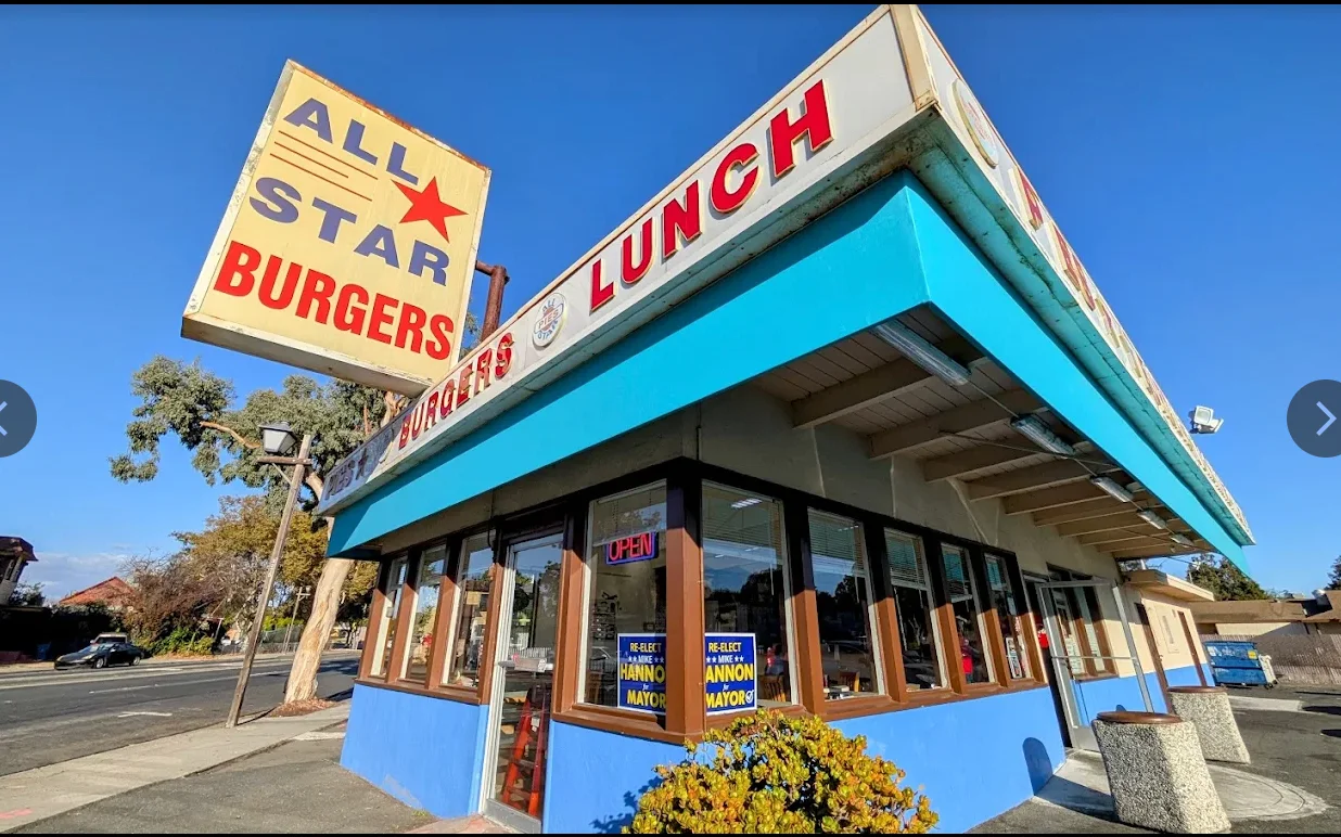 A fast food restaurant called 'All Star Burgers' featuring a bright exterior with a large sign displaying its name and offerings. The building features a mix of blue and white colors with palm trees in the background.