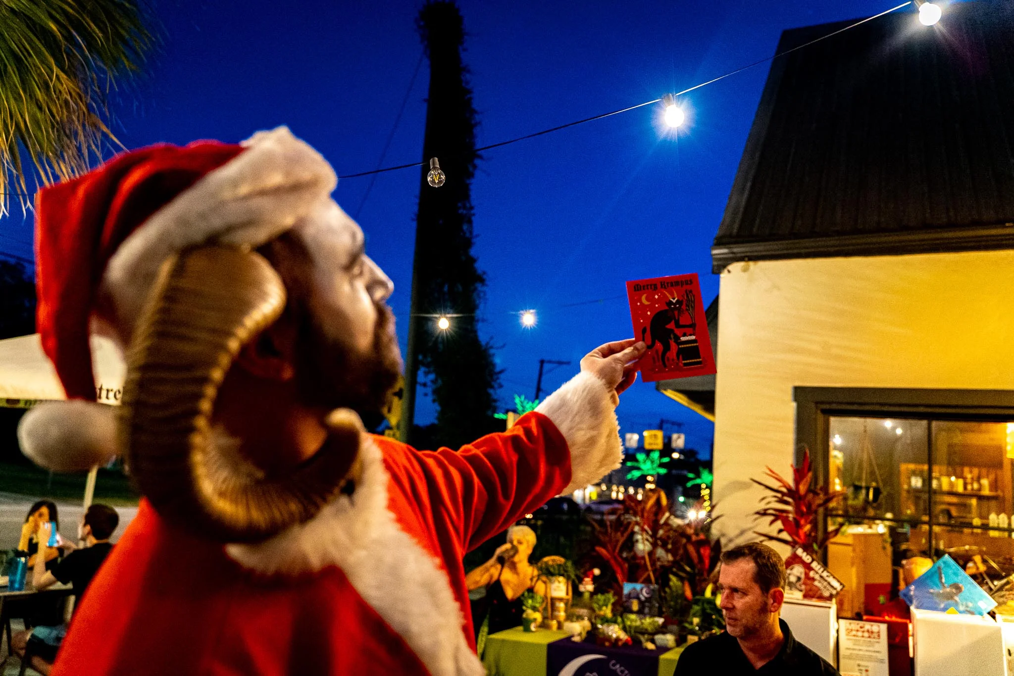 Person in festive attire holding a 'Merry Krampus' card at an outdoor event.
