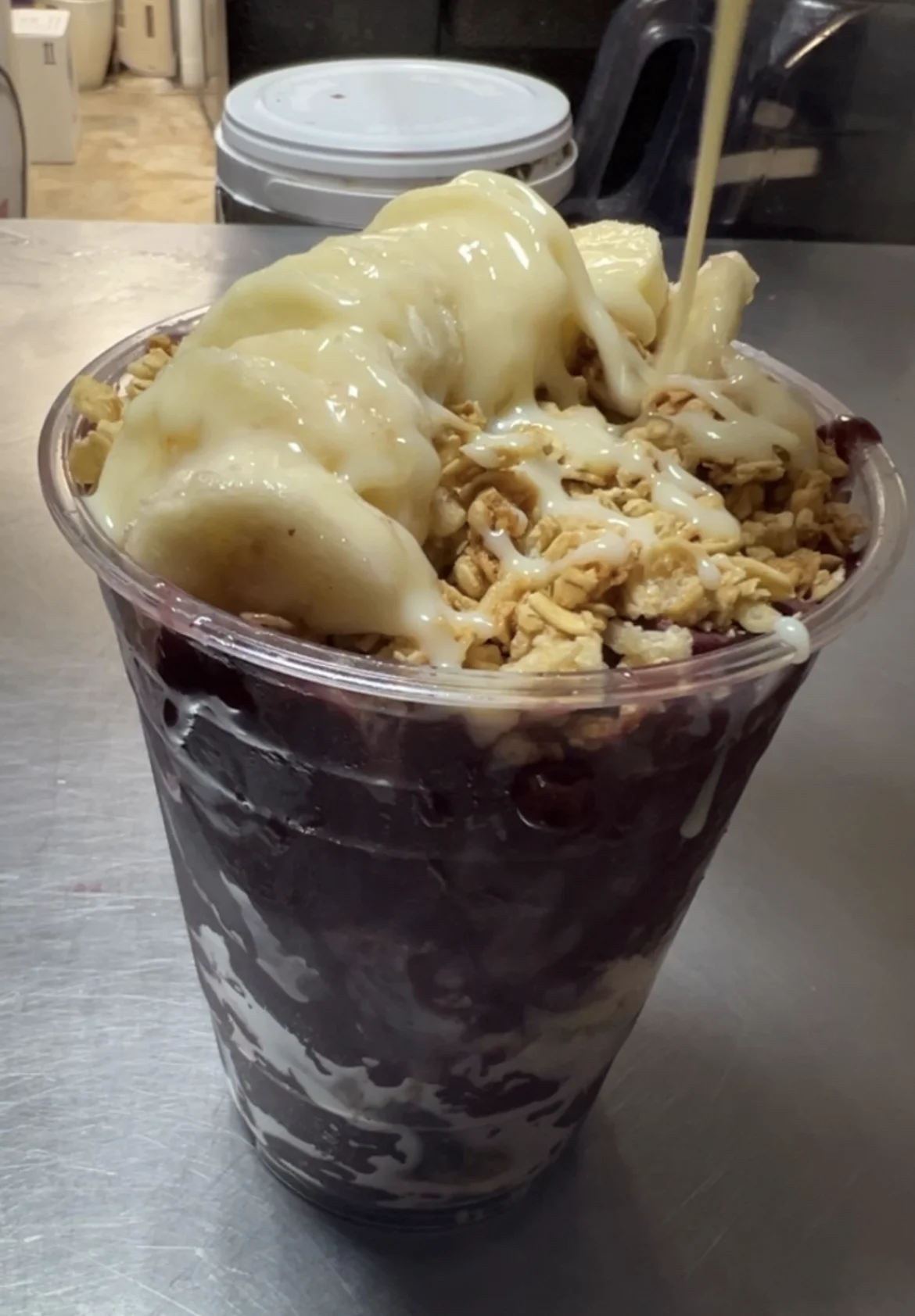 A layered dessert featuring acai, topped with banana slices, granola, and a drizzle of syrup. Served in a clear cup.