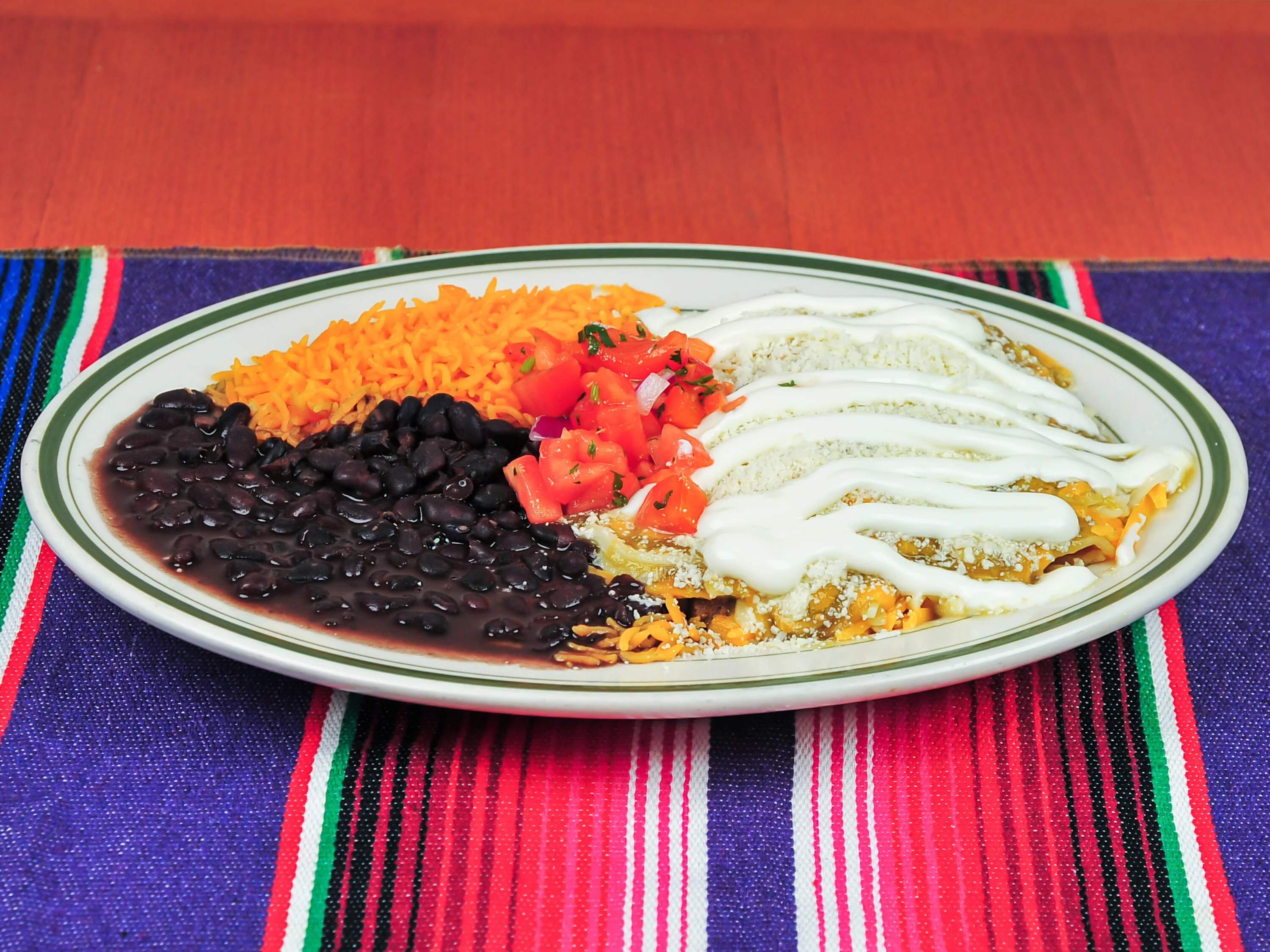 A colorful platter featuring enchiladas topped with cream, alongside black beans, Mexican rice, and diced tomatoes.