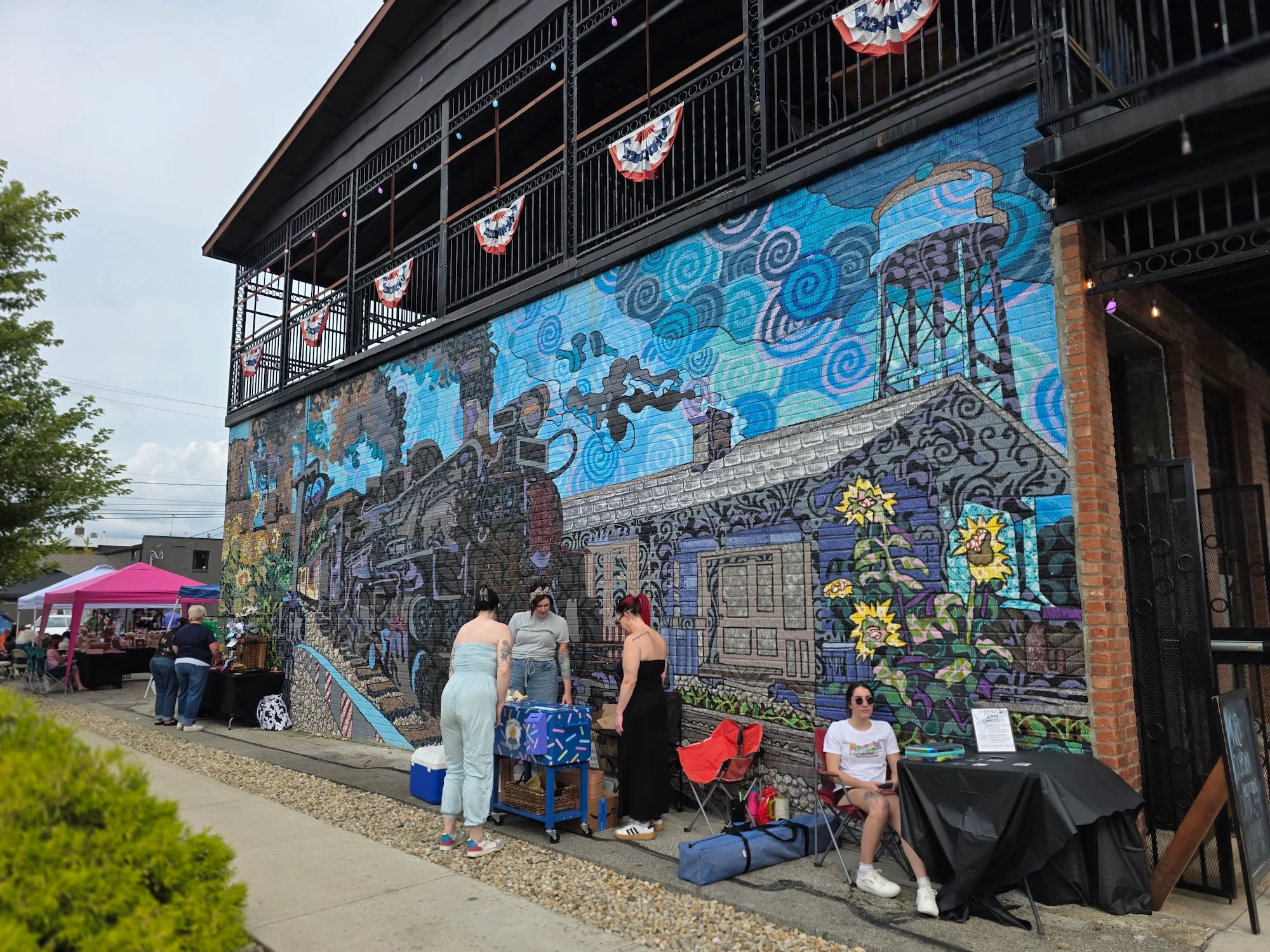 A colorful mural depicting a train and flowers on the side of a building, with people interacting at an outdoor market.