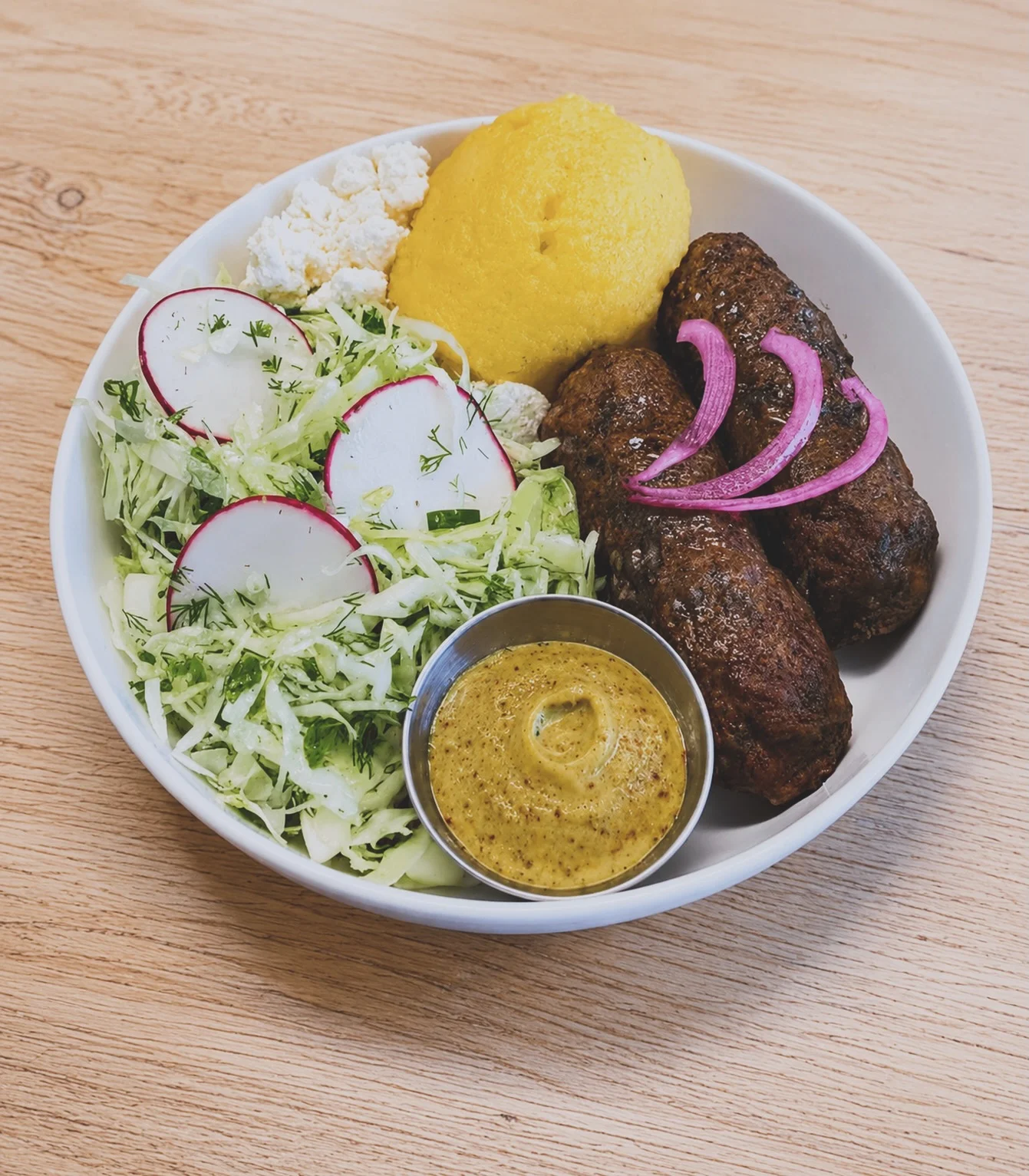 A balanced meal featuring grilled kebabs, a side of creamy polenta, fresh salad with radishes, and a mustard dip served in a bowl.
