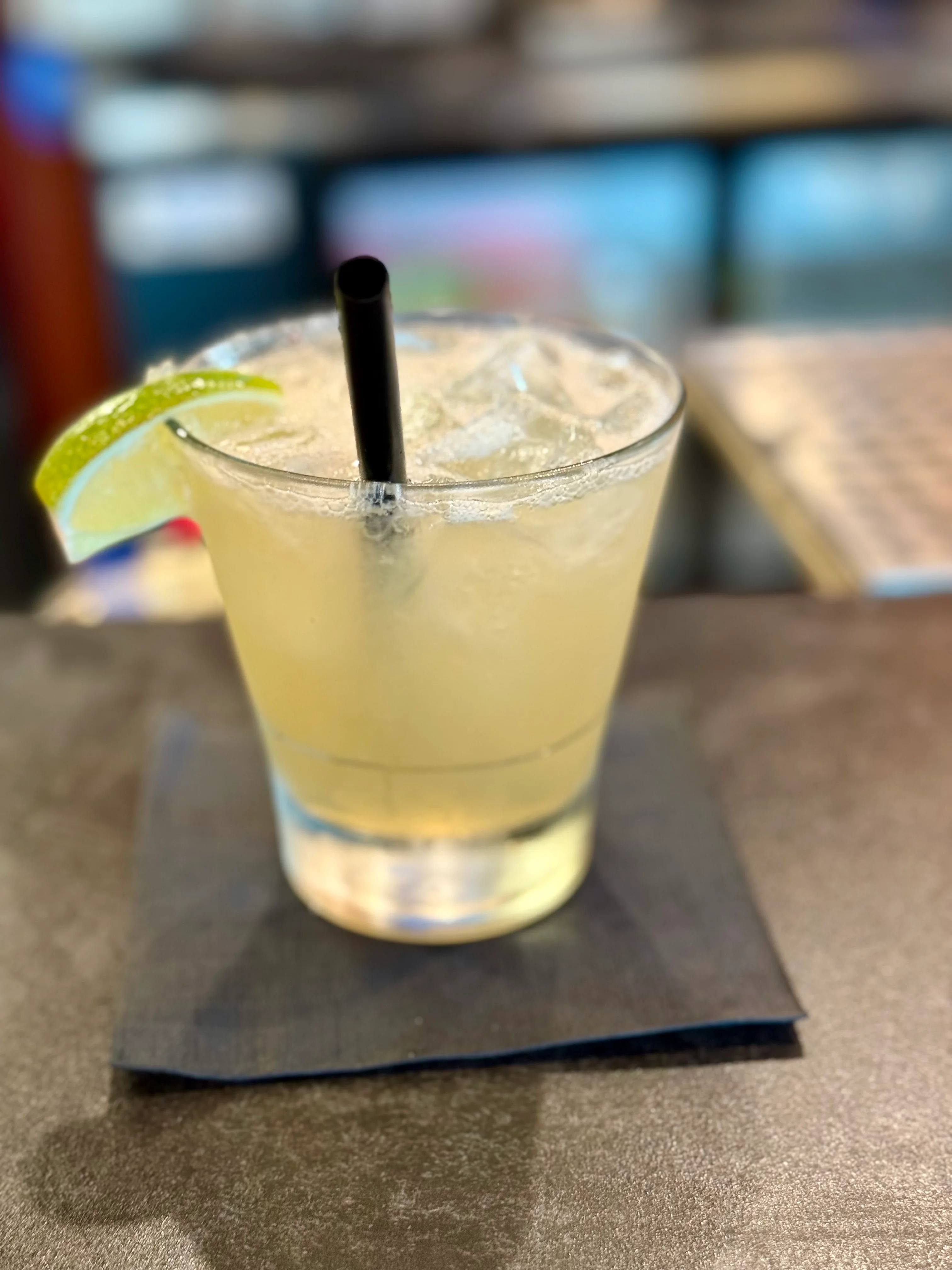 A refreshing cocktail served in a clear glass with ice and a garnish of lime. A black straw is inserted, resting in the drink.