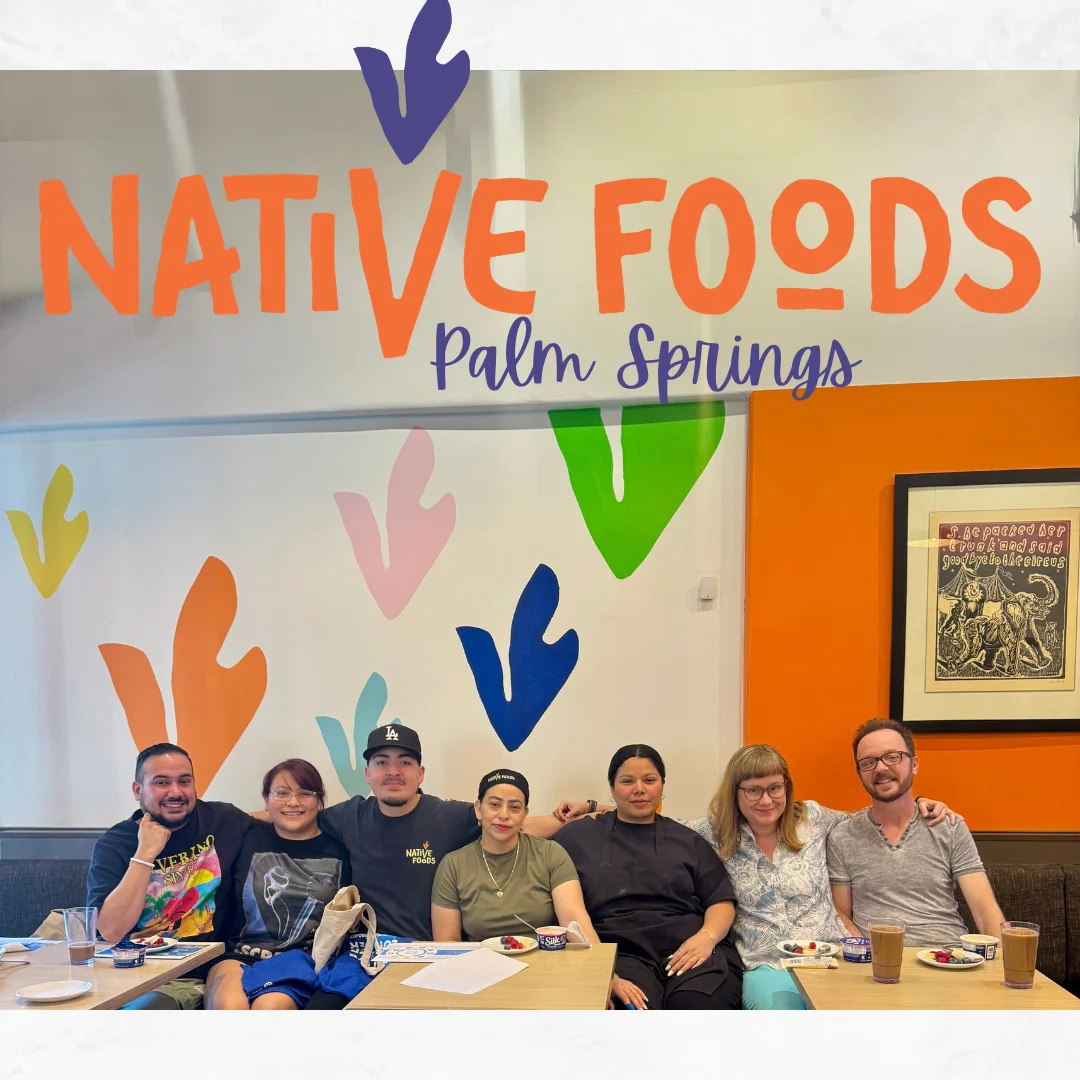A group of people enjoying a meal together in a casual dining setting, with a colorful mural in the background. The atmosphere is lively and inviting.