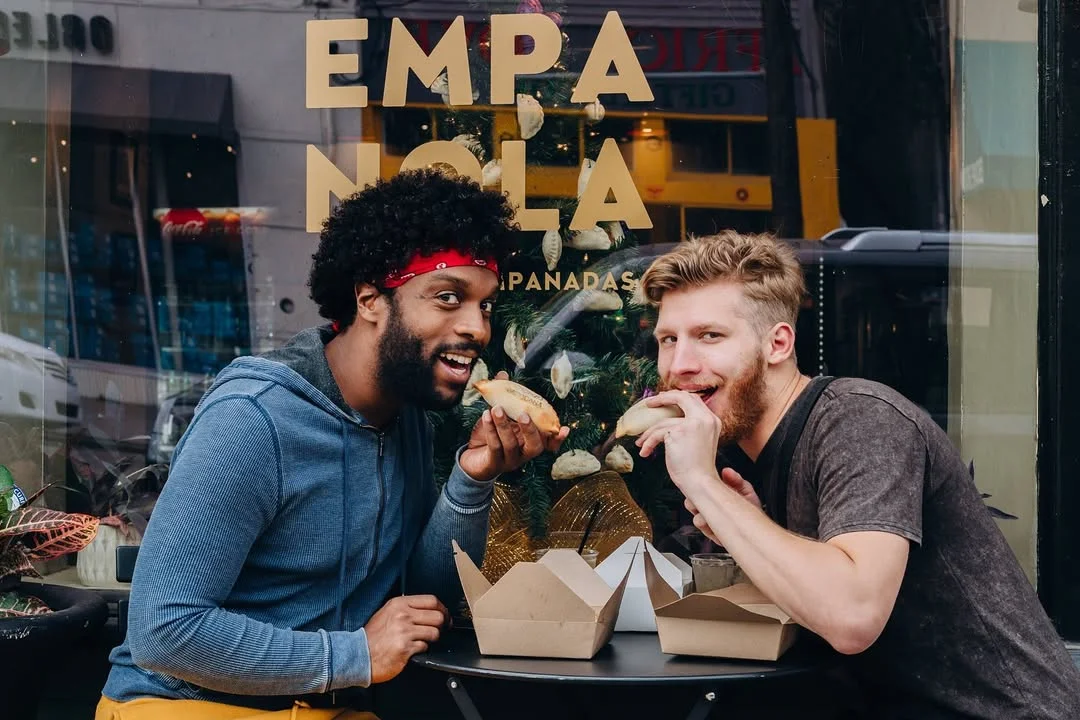 Two friends enjoying empanadas outside a vibrant restaurant