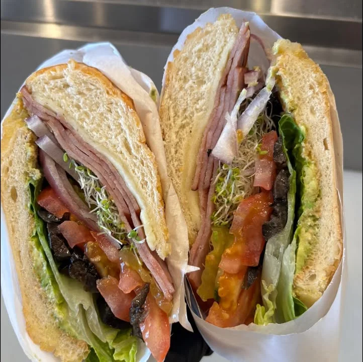 A freshly made sandwich cut in half, filled with layers of ham, vegetables, and sprouts.