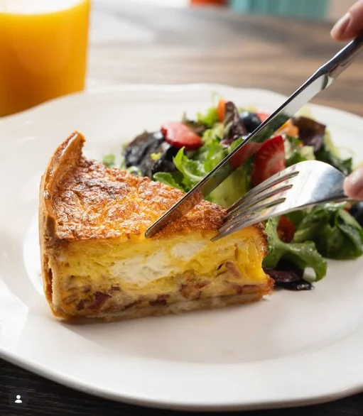 A slice of quiche served on a white plate alongside a fresh salad, with a fork cutting into the quiche.
