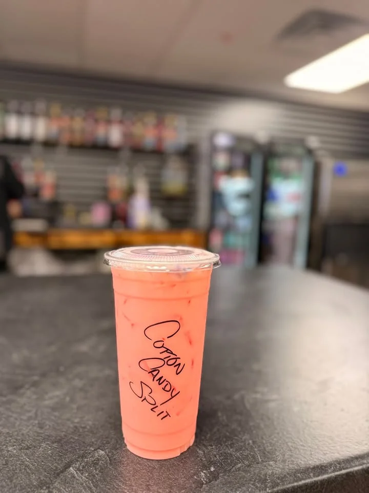 A clear plastic cup filled with a pink beverage, sitting on a dark countertop. The cup is labeled 'Cotton Candy Split' and contains ice.