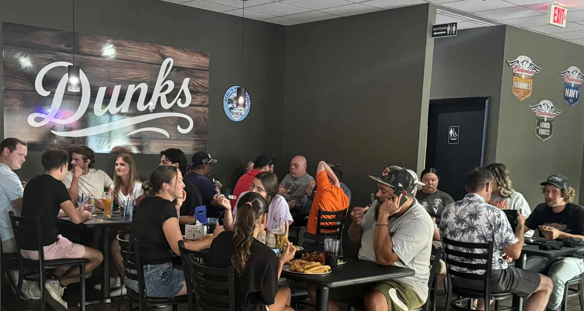 Groups of customers dining inside Dunks Bar & Grill, sitting at tables with food and drinks, under a large wooden wall sign that reads “Dunks.”
