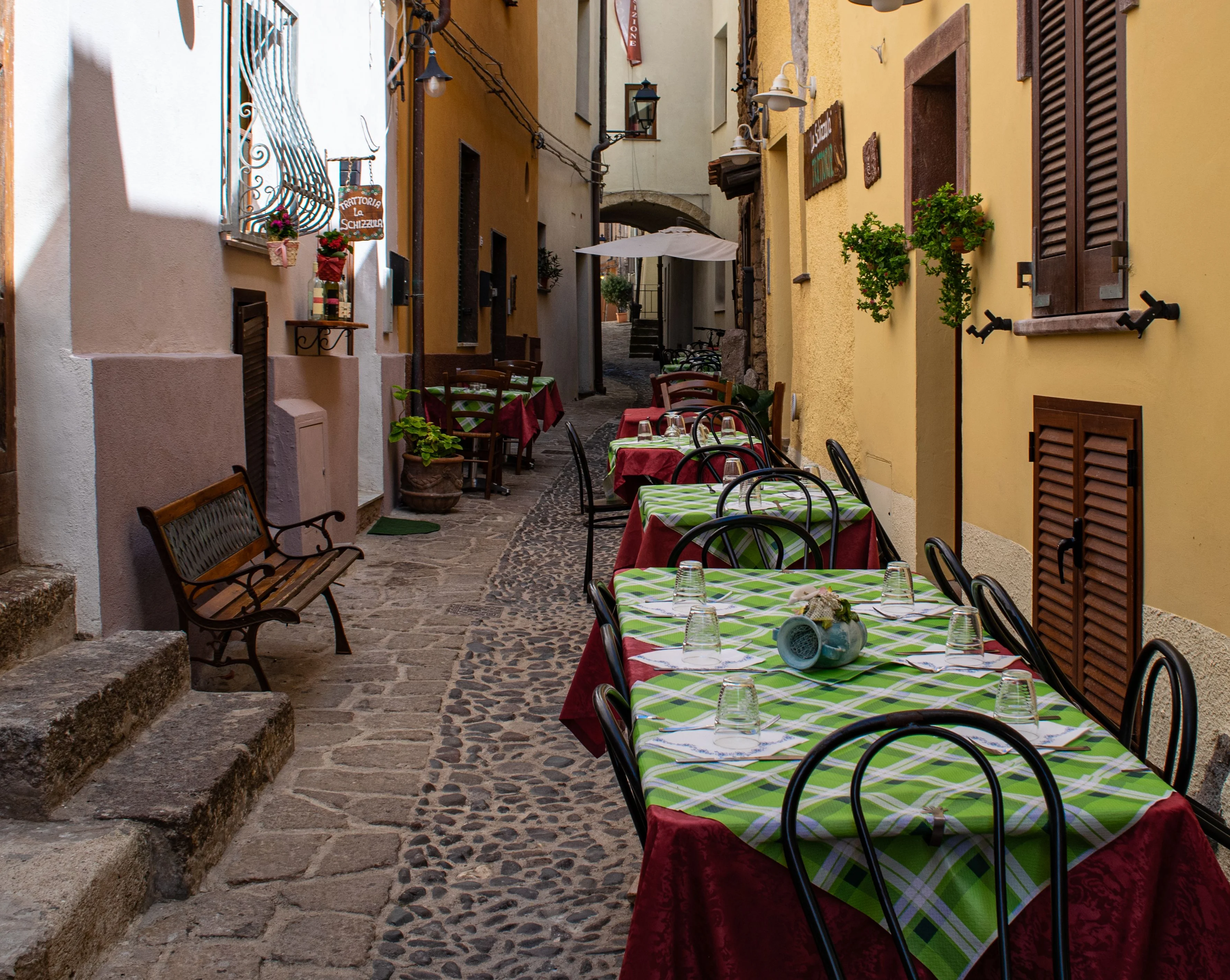 Sunlit Roman cobblestone alley with outdoor dining tables and colorful buildings, reflecting the hospitality behind Daje Pinsa Roman Pizza in Orlando.
