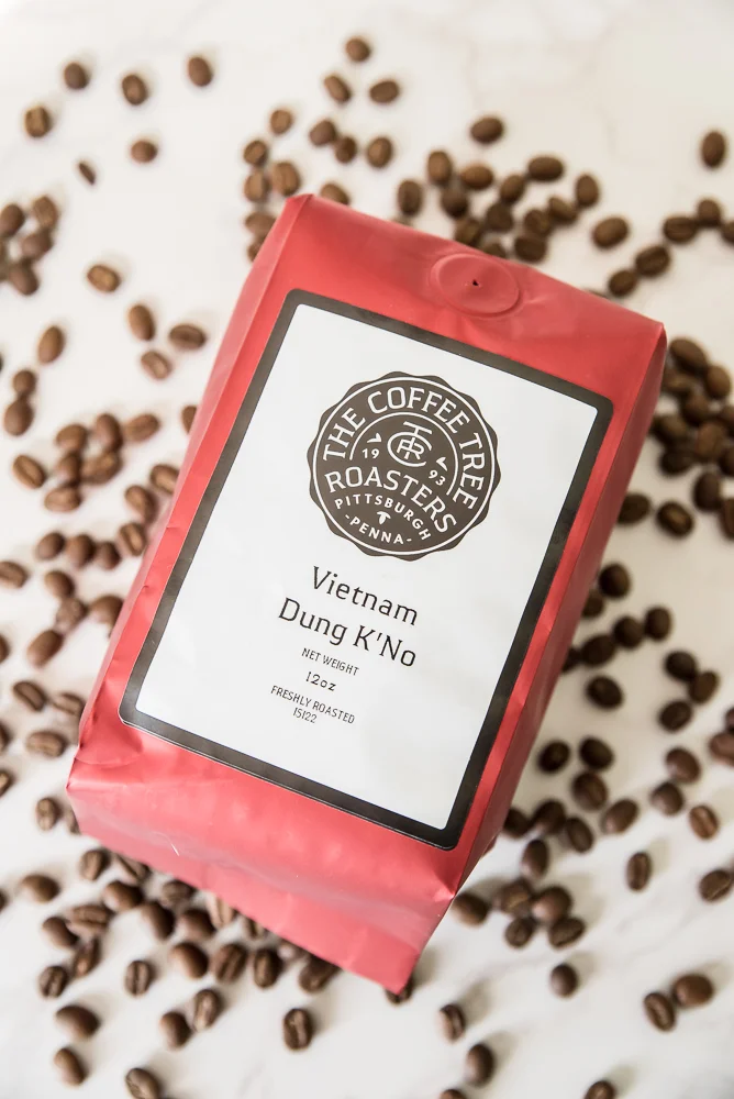 Red coffee bag labeled "Vietnam Dung K'No" from The Coffee Tree Roasters is surrounded by scattered coffee beans on a marble surface.
