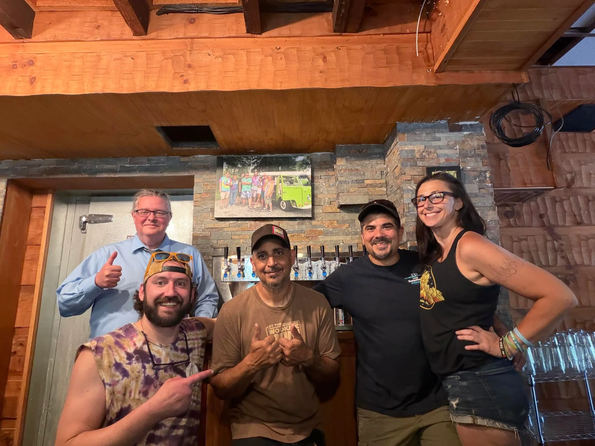 A group of five people posing together at a bar, with a picturesque background of wooden decor and a wall artwork.