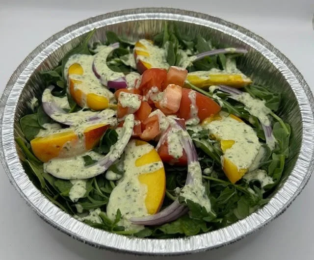A fresh salad featuring arugula, tomatoes, yellow peaches, and red onions, topped with a creamy dressing in a silver serving dish.