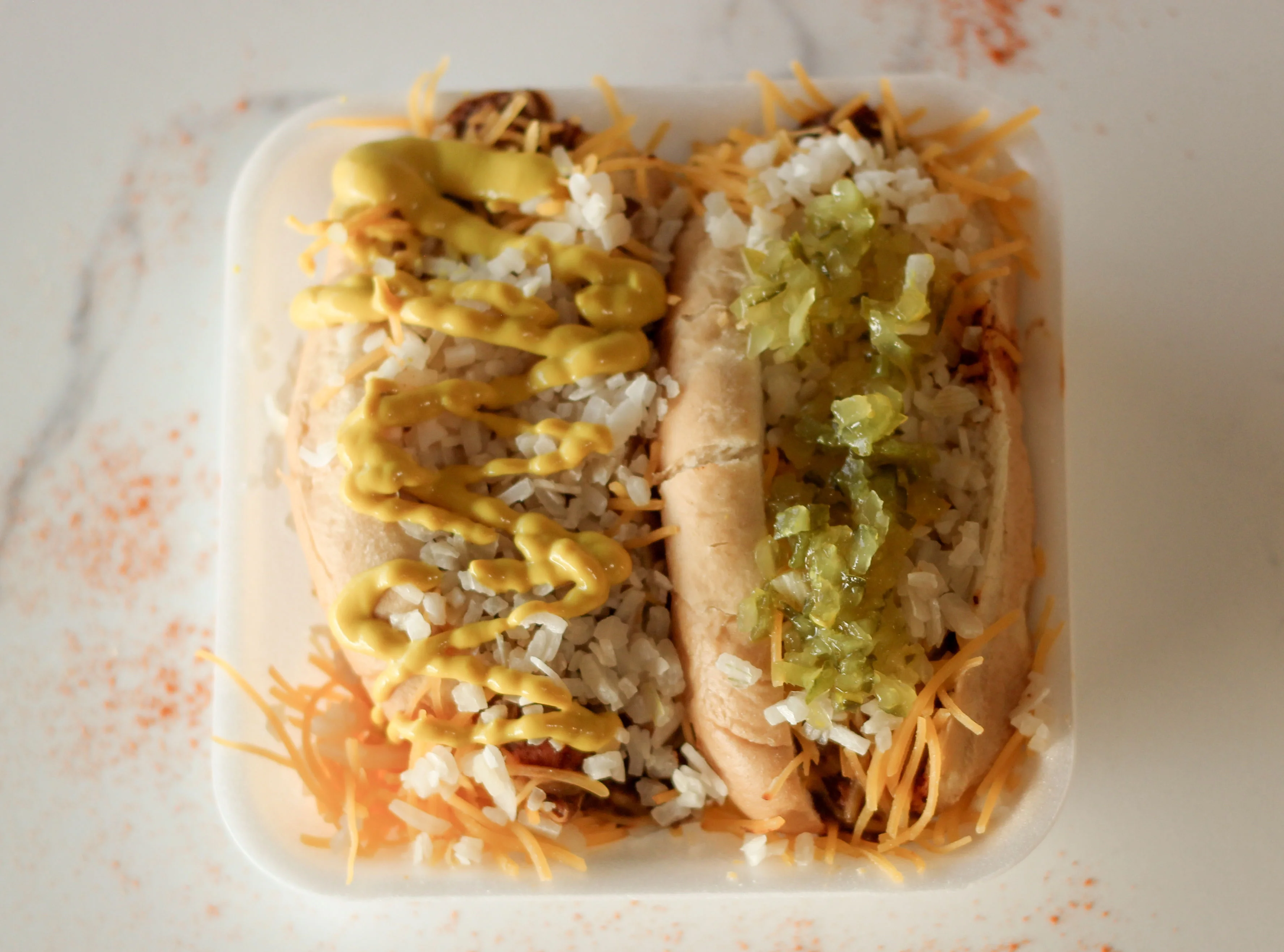 A container with two hot dogs topped with mustard, relish, and cheese, surrounded by rice, served on a white background.
