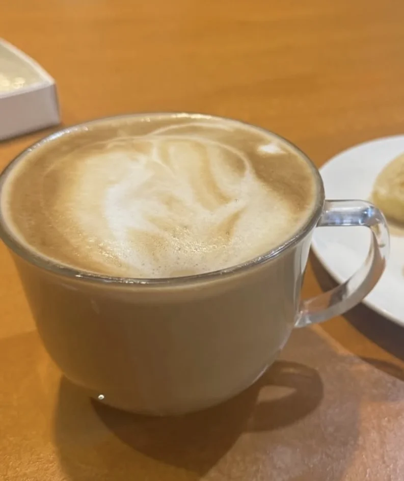 A cup of creamy latte with a swirled foam on top, placed on a wooden table next to a plate with baked goods.