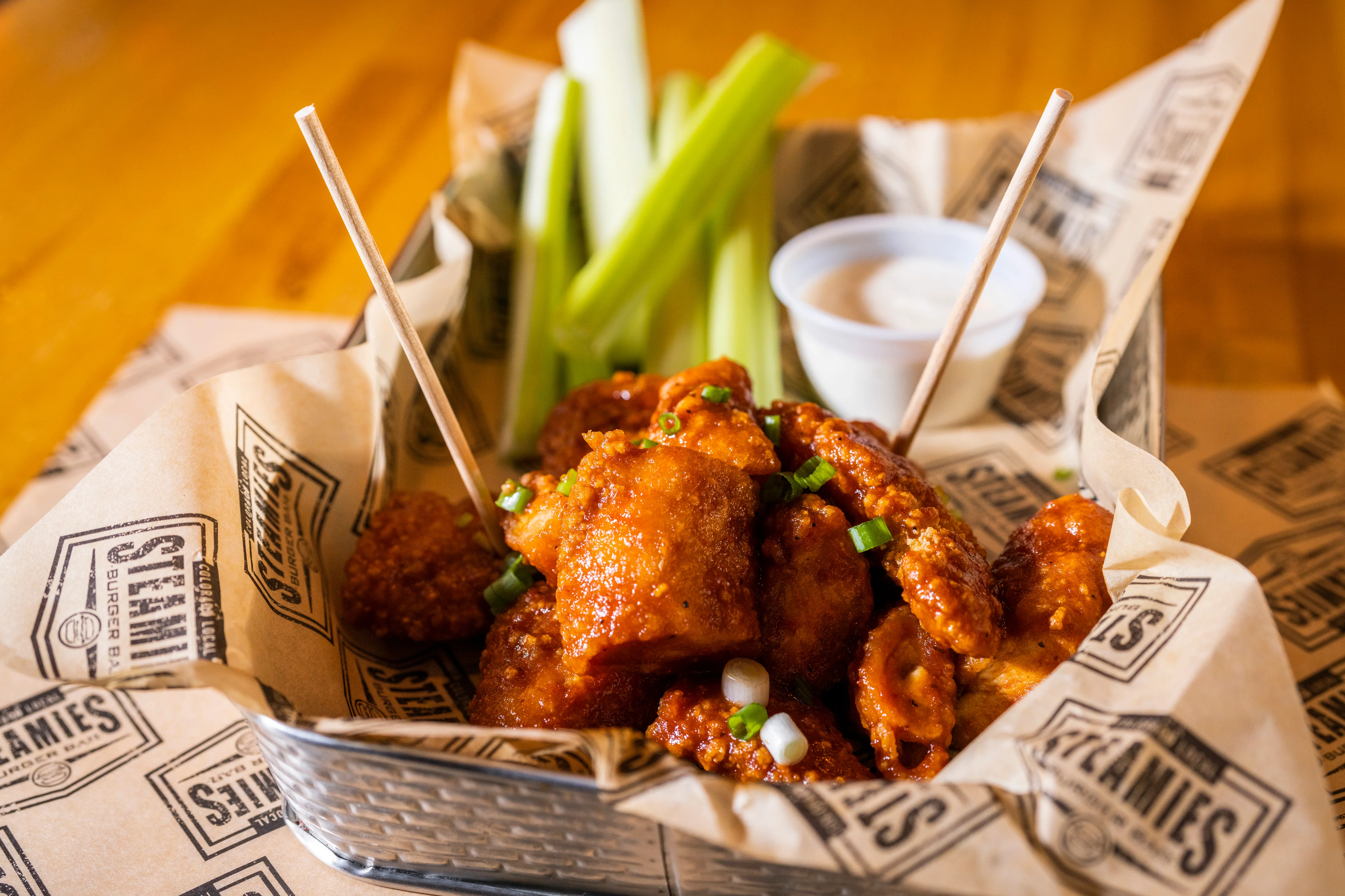 A serving of spicy chicken wings accompanied by celery sticks and a small cup of ranch dressing, presented in a basket lined with paper.