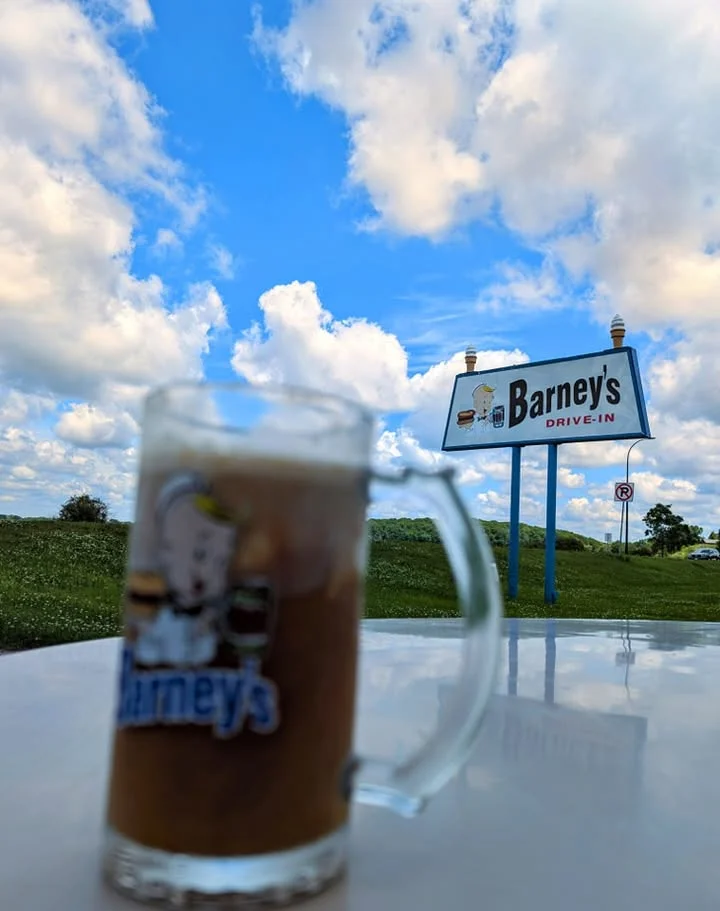 A mug of cold beverage is set against a blue sky and a roadside sign of a drive-in restaurant.