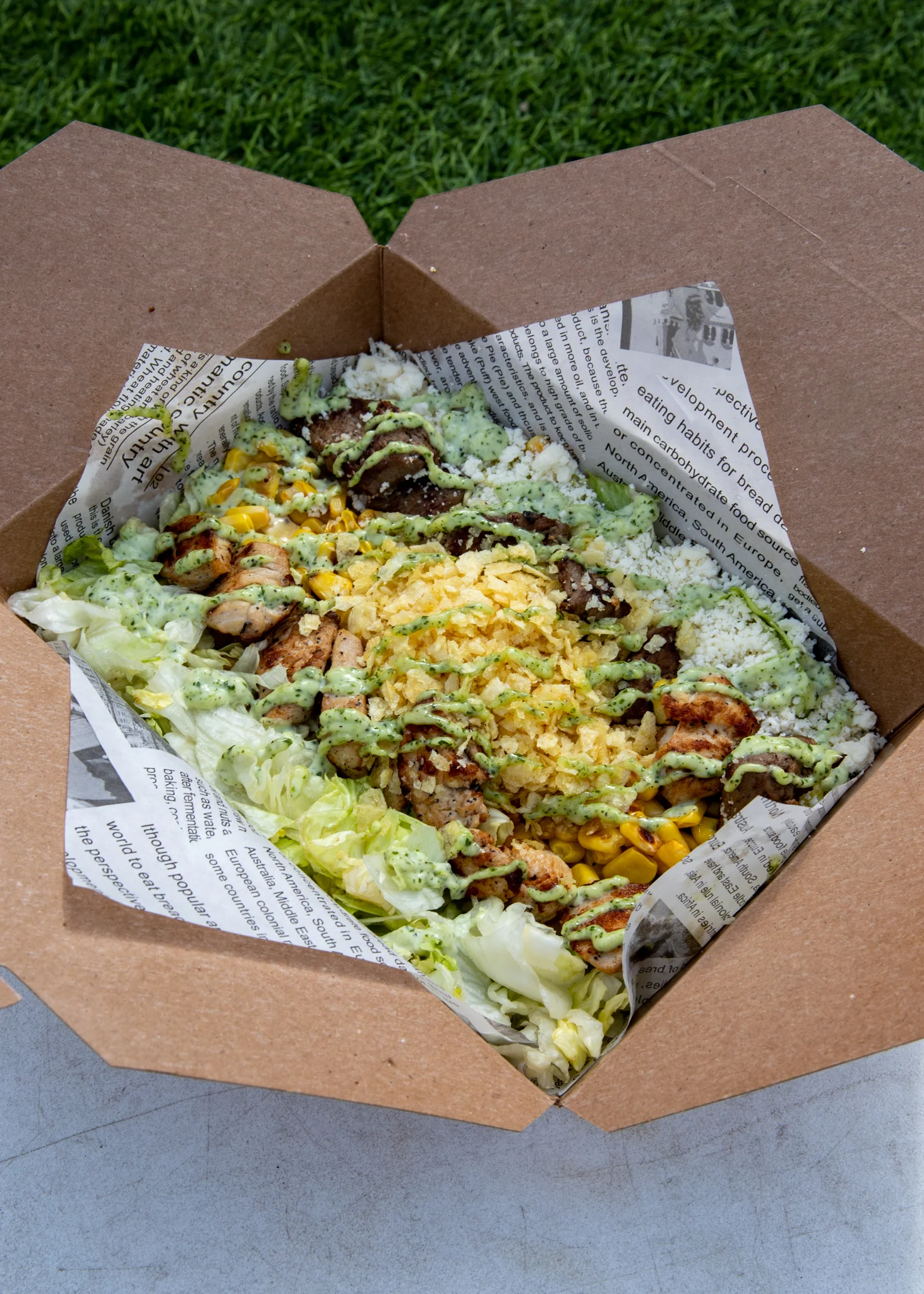 A takeout box filled with a mix of grilled meats, rice, lettuce, corn, and drizzled with green sauce.