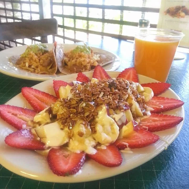 A colorful dessert featuring sliced strawberries, yogurt drizzled with honey, granola, and a glass of orange juice, with tacos in the background.