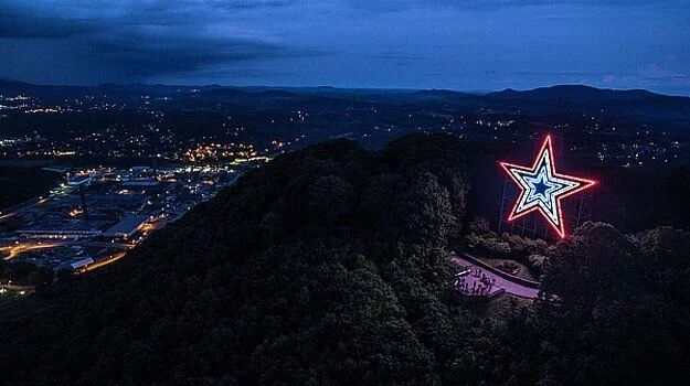 A large, illuminated star on a hillside, glowing against a twilight sky. Below, a small gathering of people can be seen near a pathway while the city lights twinkle in the background.