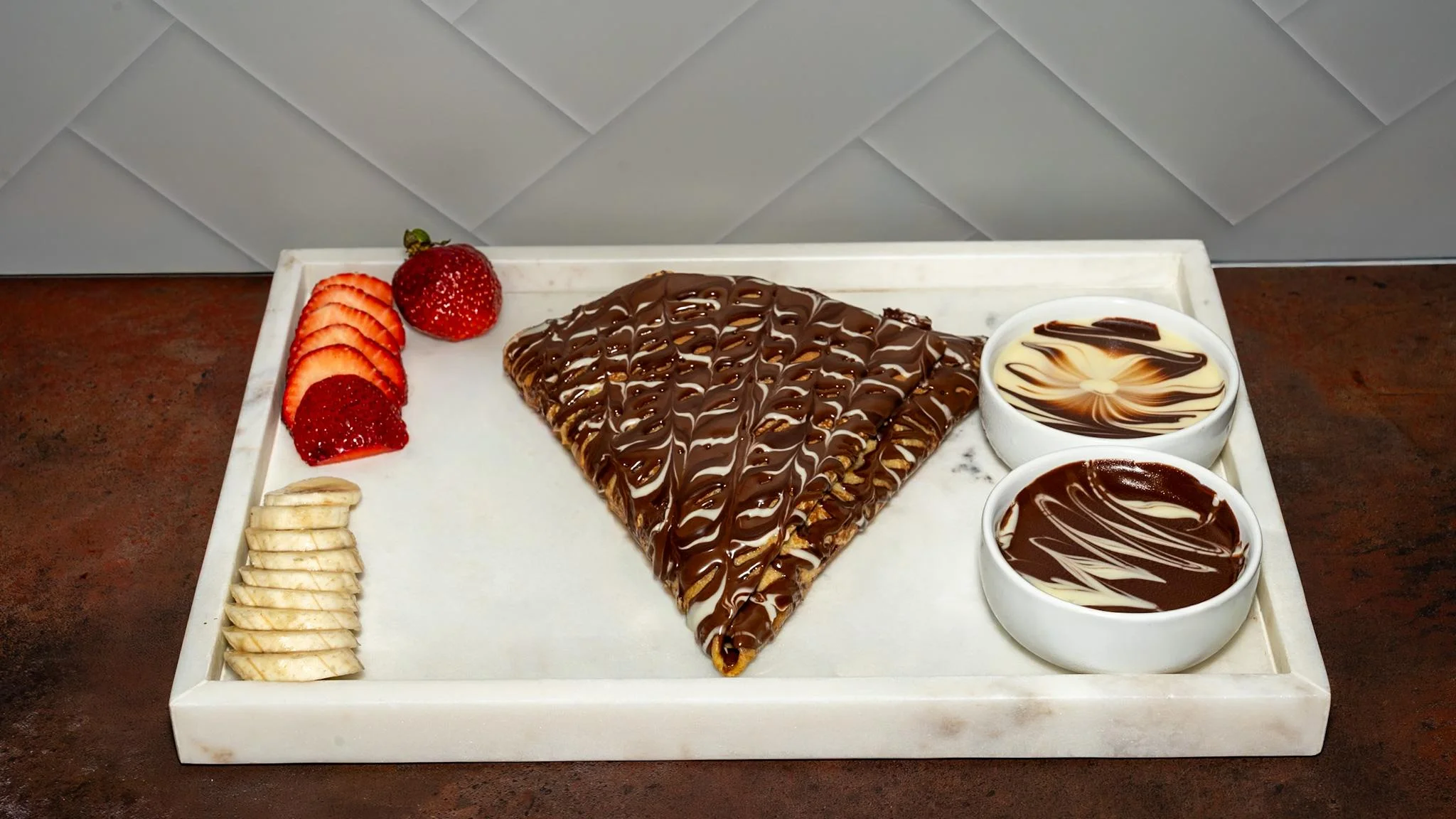 A chocolate-filled crepe drizzled with chocolate and served with strawberries and banana slices, alongside creamy dipping sauces.