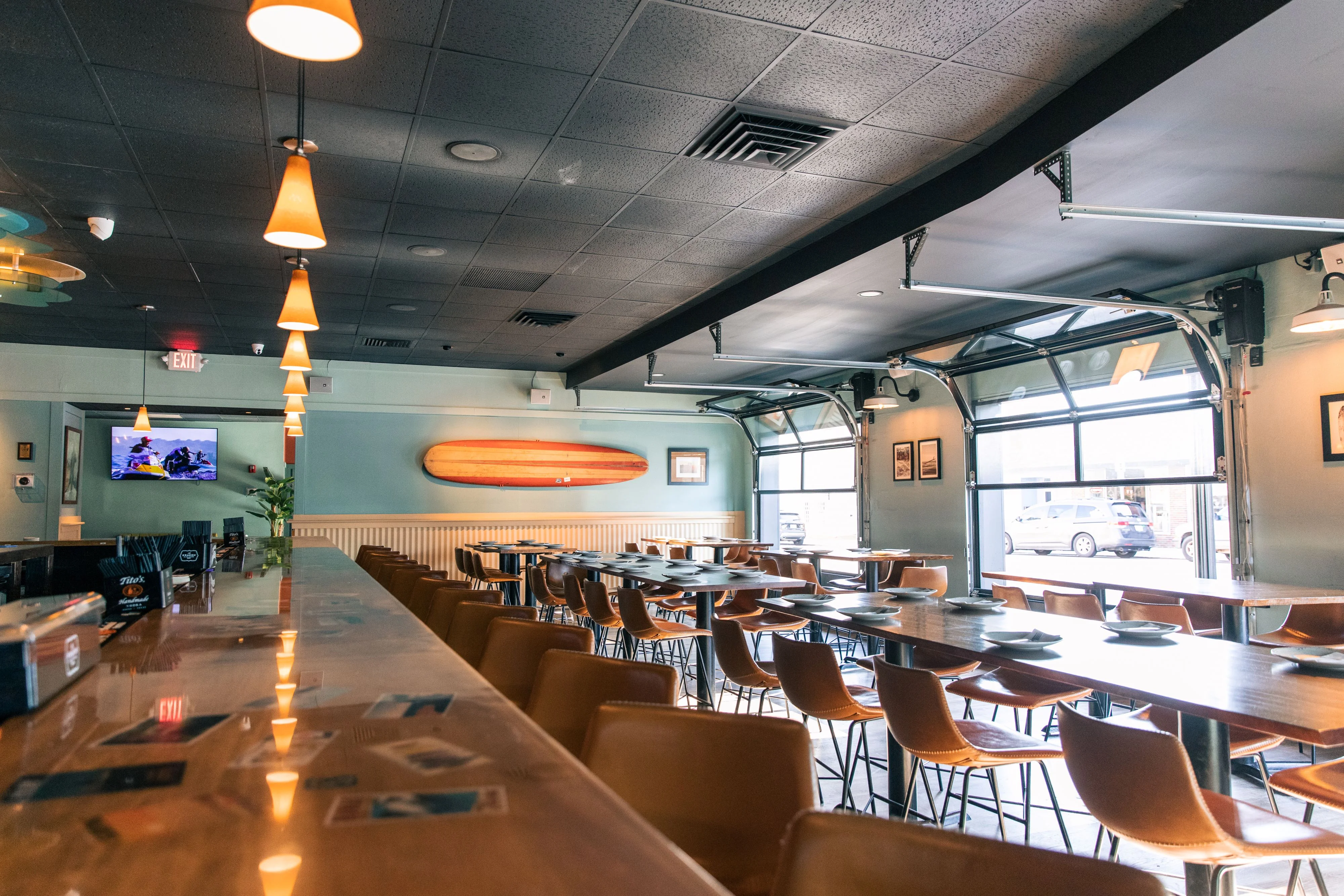 A spacious restaurant interior featuring long wooden tables and leather chairs, with a surfboard displayed on the wall and large garage-style windows letting in natural light.