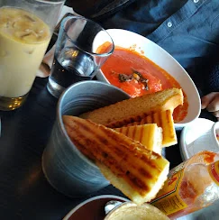 A bowl of tomato soup served alongside grilled cheese sandwiches and a cold drink, all on a table.