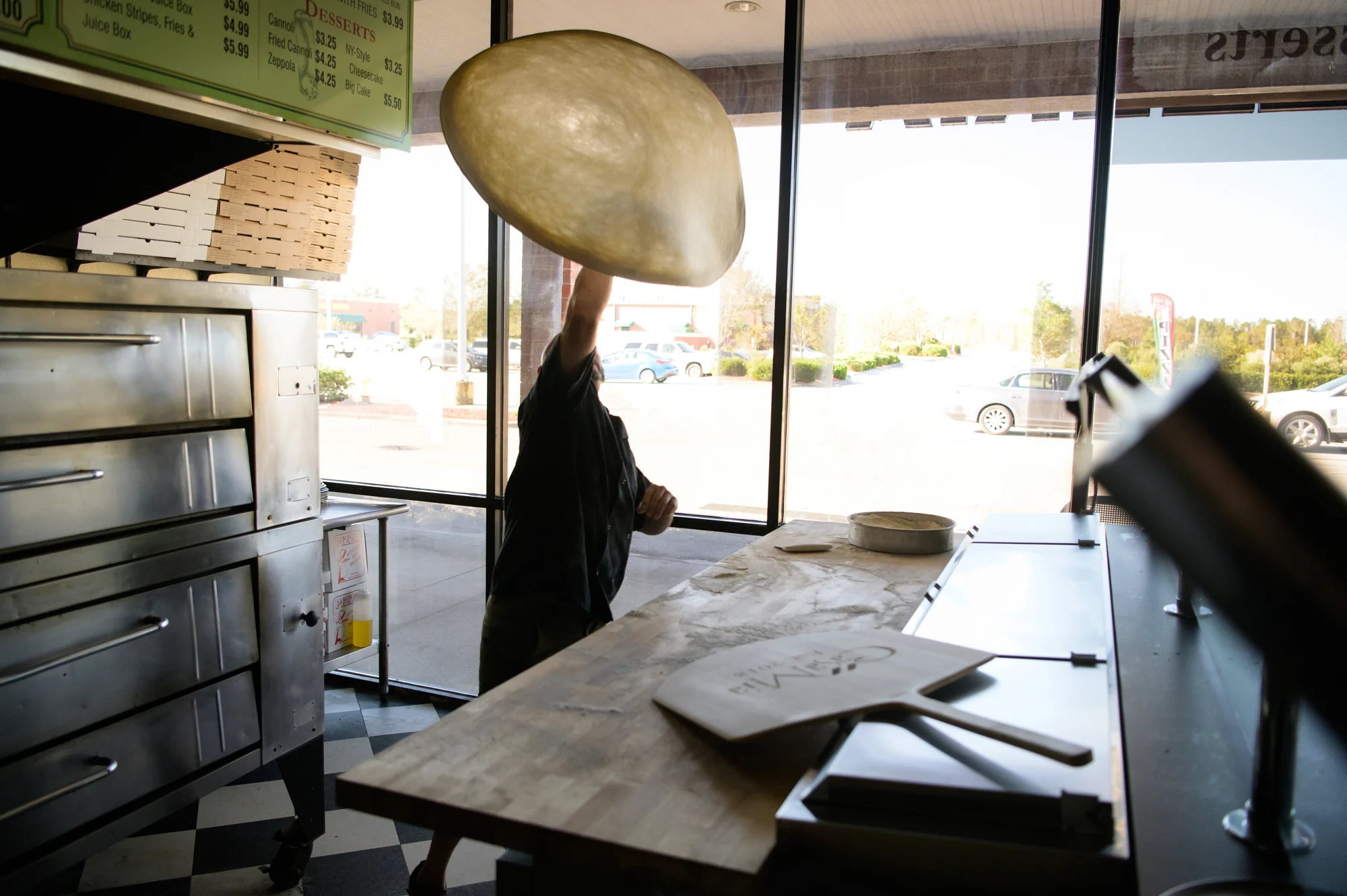 A chef skillfully tossing pizza dough in a pizzeria with a view of a sunny day outside.