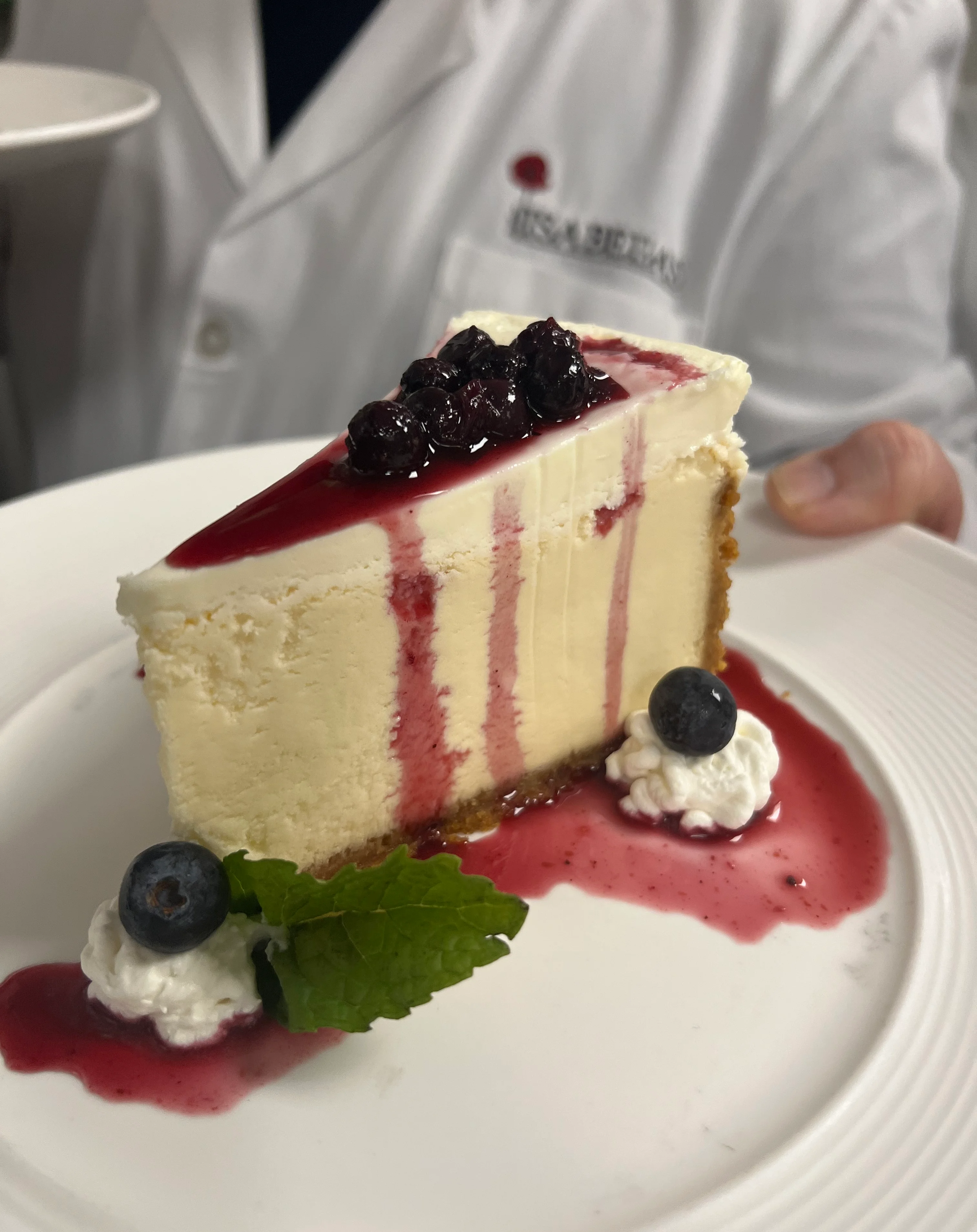 A slice of creamy cheesecake topped with blueberry compote and drizzled with berry sauce, served on a white plate with dollops of whipped cream, fresh blueberries, and a mint leaf garnish. A person in a white coat with the name “ISABELLA’S” is holding the plate.