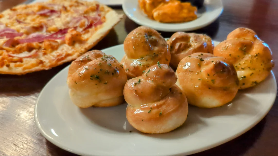 Butter-covered made-from-scratch garlic knots with BBQ chicken and buffalo wings in the background.