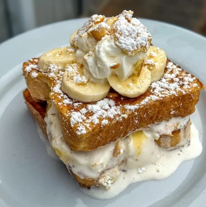 A sweet dish of stacked French toast filled with cream and topped with banana slices and whipped cream, dusted with powdered sugar.