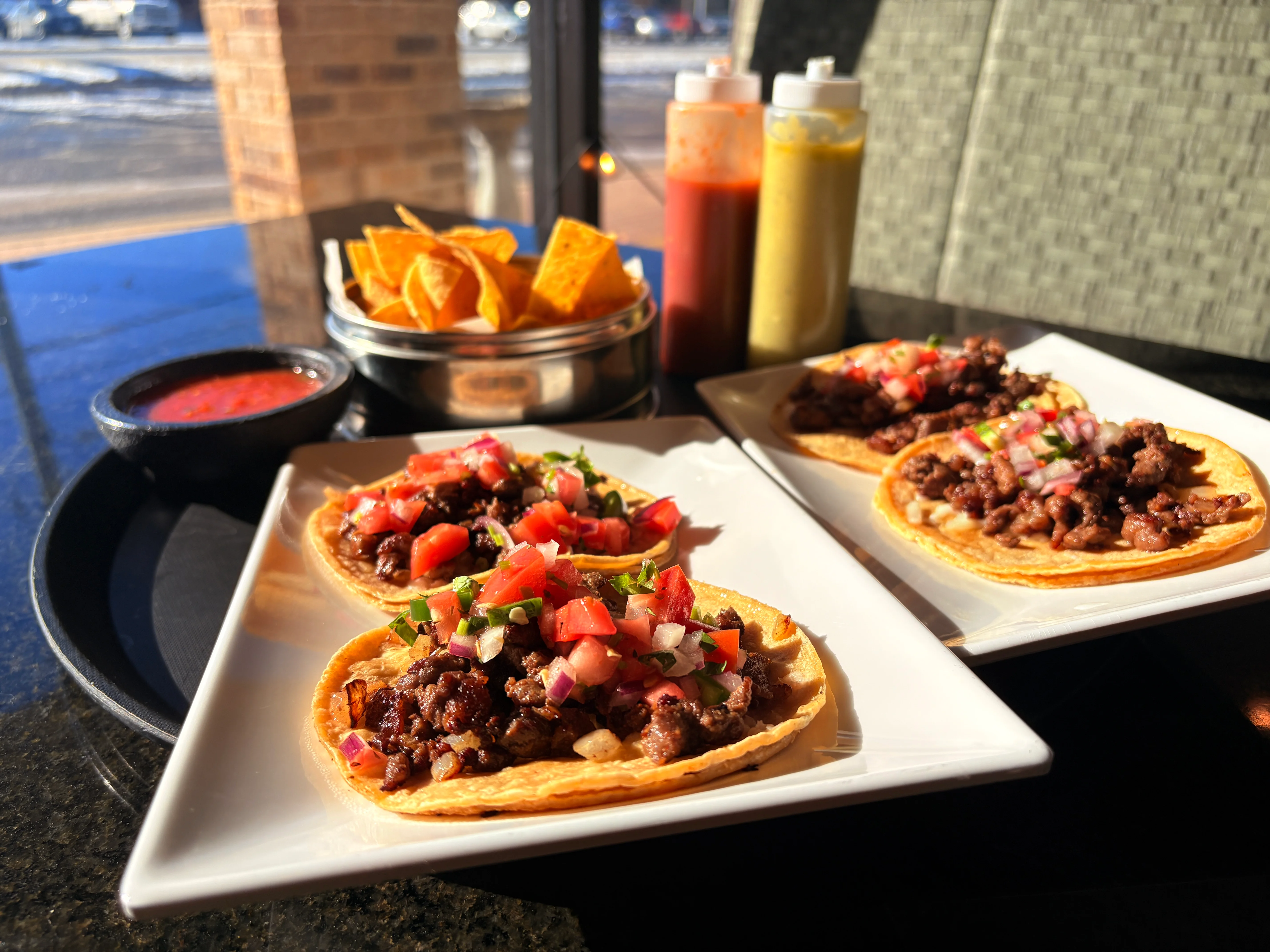 A sunny dining scene featuring tacos filled with seasoned meat and fresh vegetables, accompanied by tortilla chips and a variety of sauces.