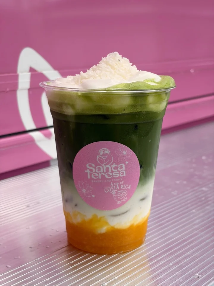 A vibrant layered beverage in a clear cup, showcasing green matcha, creamy white, and orange layers topped with coconut flakes.