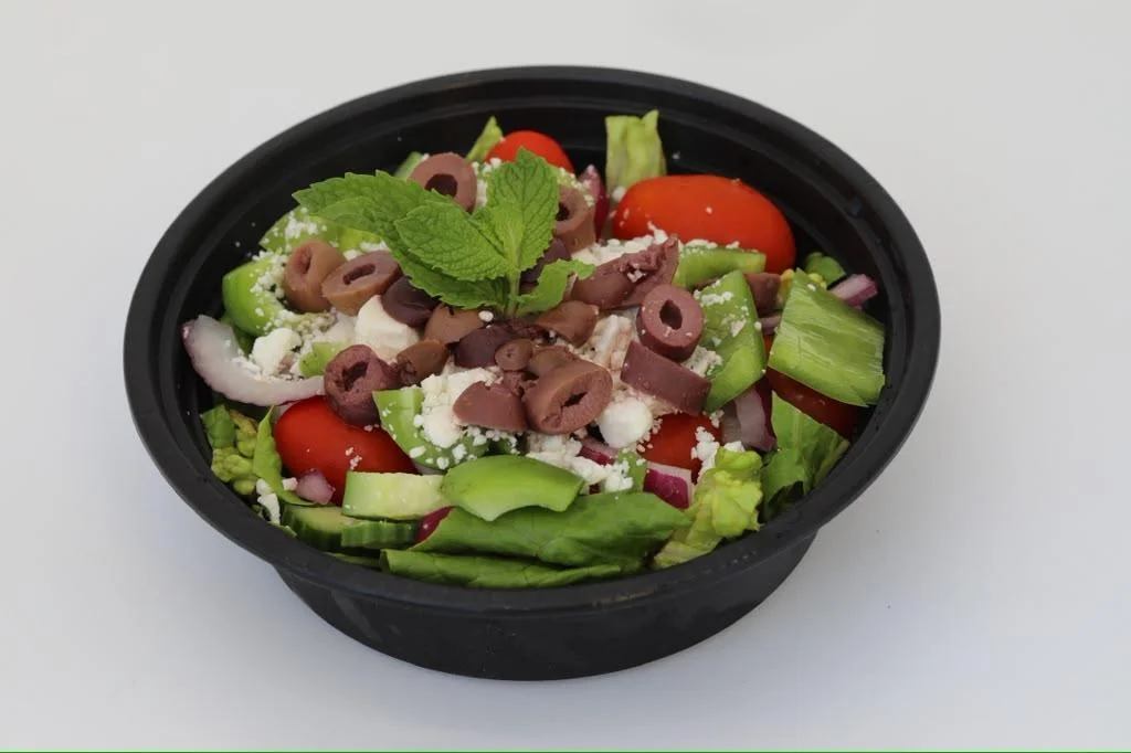 A fresh salad in a black bowl with assorted vegetables, olives, and a mint leaf garnish.