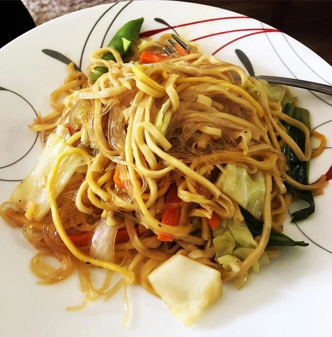 A plate of stir-fried noodles mixed with assorted vegetables including cabbage, carrots, and snap peas. The dish features a mix of thin rice noodles and egg noodles.