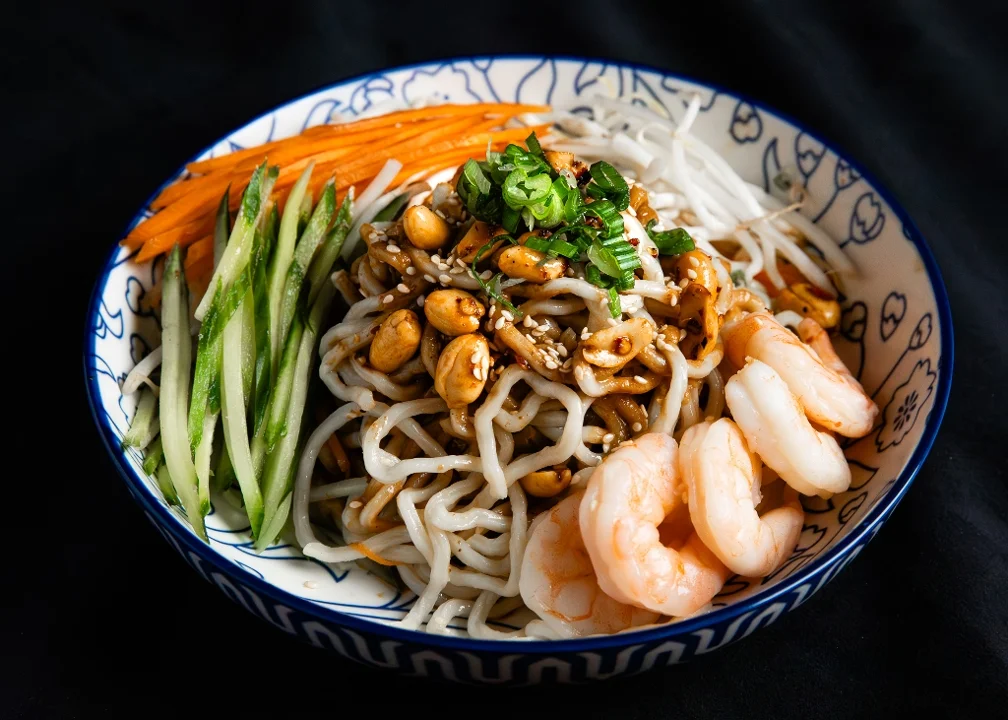 A bowl of noodles topped with shrimp, peanuts, and vegetables, including carrots and cucumbers, with sesame seeds and green onions.