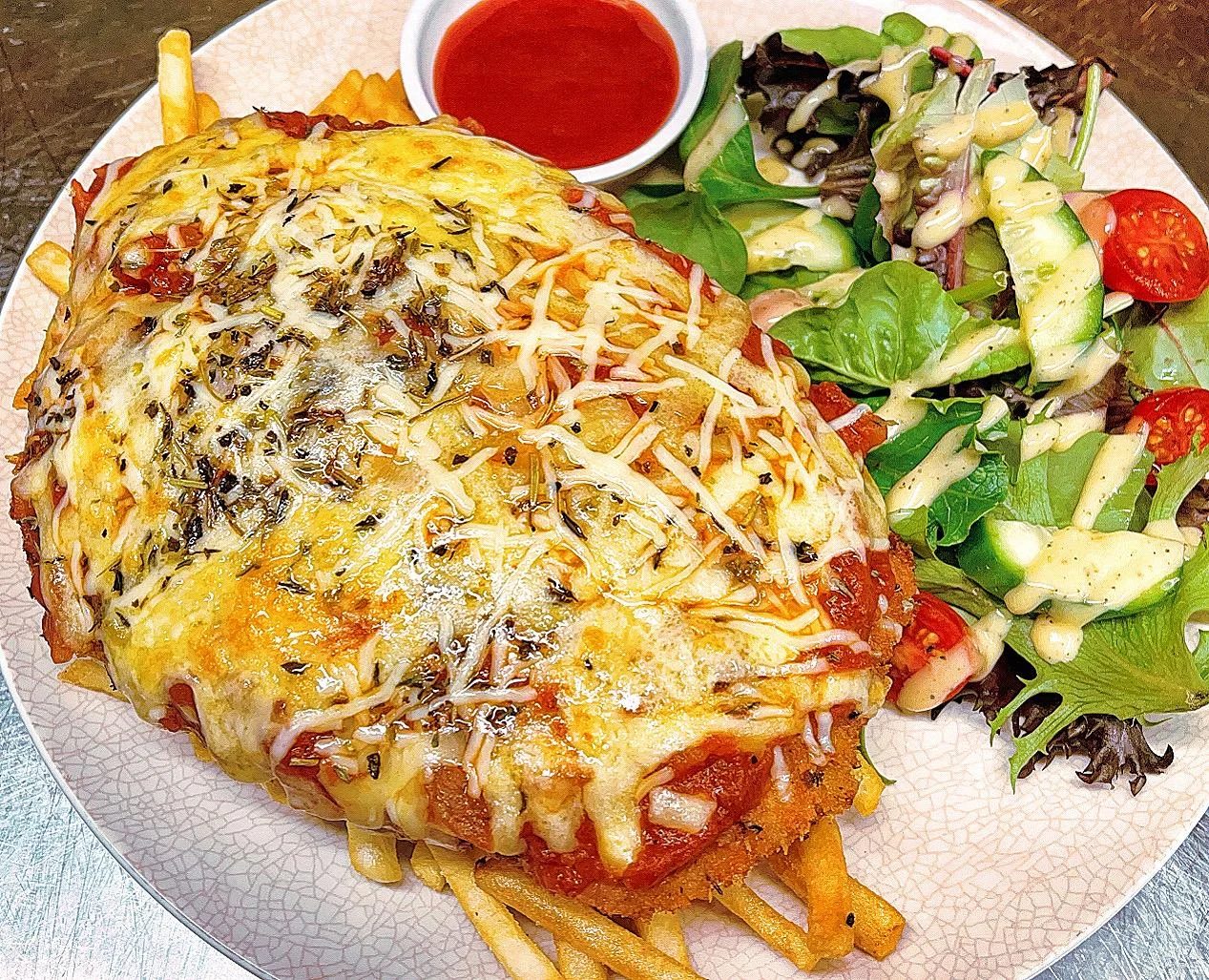 A loaded dish featuring a breaded item topped with cheese and sauce, served alongside fries and a fresh salad with dressing.