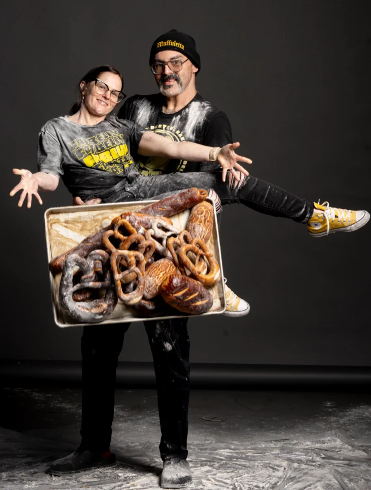 A couple covered in flour posing playfully while holding a tray full of pretzels.